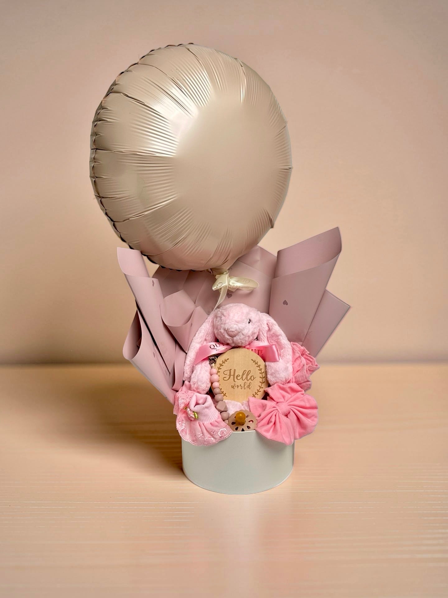 Blush pink baby girl gift bouquet box featuring champagne gold foil balloon, plush pink bunny toy, wooden milestone disc, pink baby booties, headband, and pacifier clip by Qute Gifts Doha Qatar