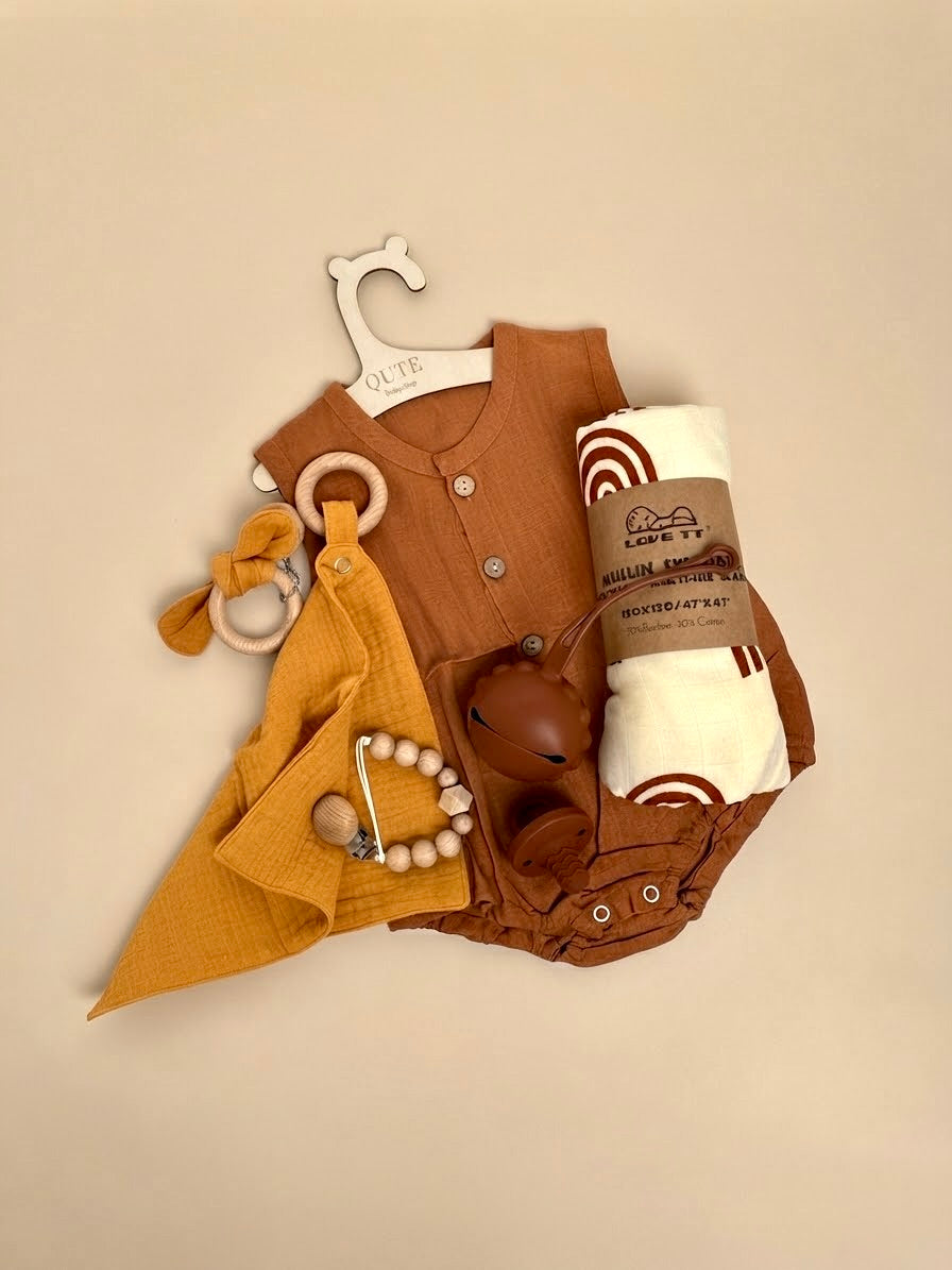 Luxury newborn baby gift set in a white keepsake suitcase by Qute Qatar, featuring a 3-6 months rust and mustard outfit, rainbow muslin swaddle, pacifiers, and wooden accessories for delivery in Doha.