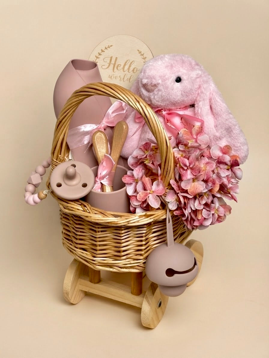 Luxury newborn baby girl feeding gift set in a wheeled wooden carriage basket by Qute Qatar, featuring a pink silicone feeding set, plush bunny, milestone disc, and floral accents for delivery in Doha.