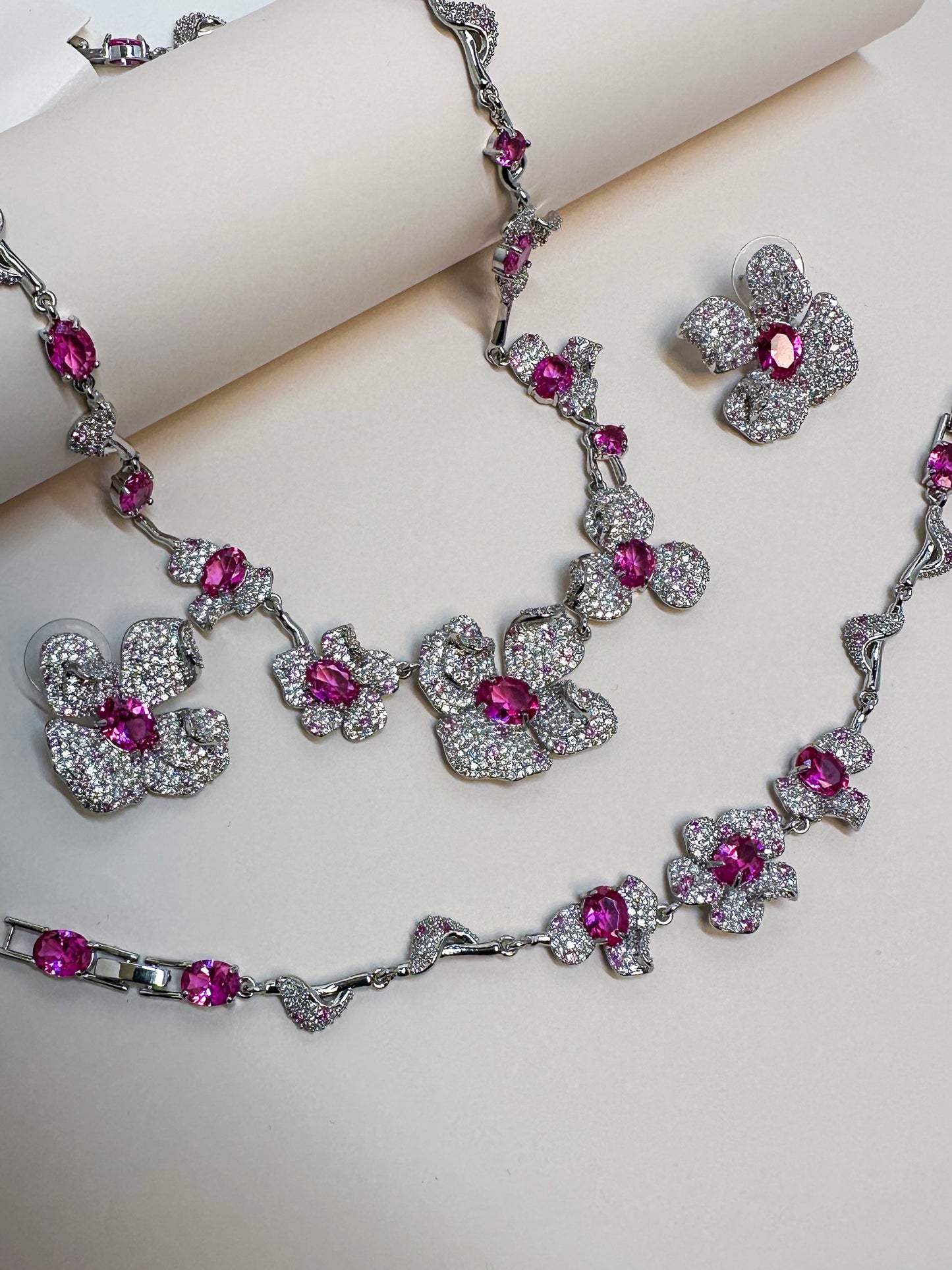 Elara jewelry set displayed on dark velvet featuring a silver-plated floral statement necklace with brilliant fuchsia pink crystal centers and heavy pave-encrusted petals, a matching link bracelet, and coordinating stud earrings in Doha Qatar