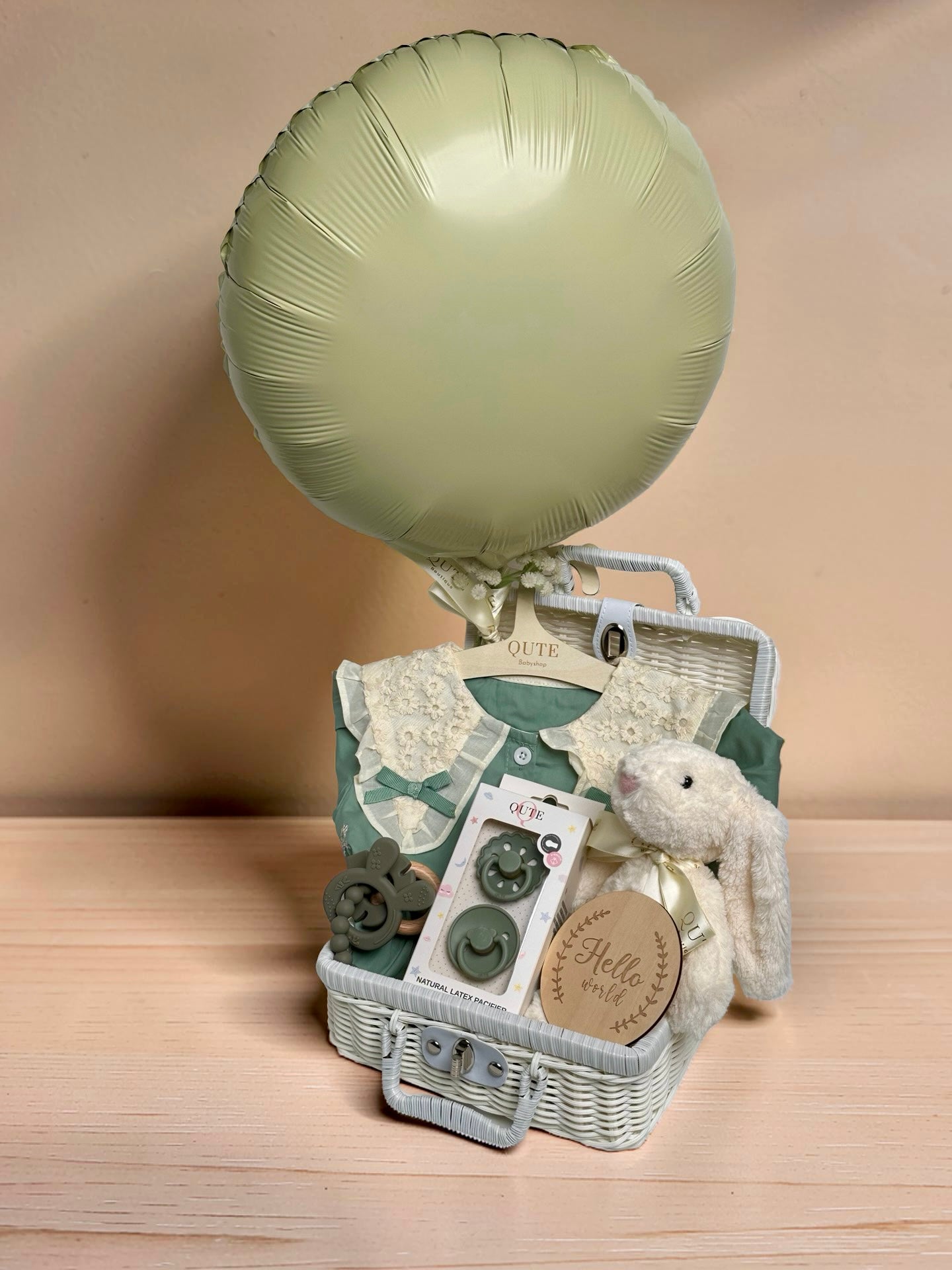 Deluxe sage green baby girl gift set in white woven suitcase hamper with lace collar dress, Qute natural latex pacifiers, silicone teether ring, plush bunny toy, and wooden milestone disc by Qute Gifts Doha Qatar