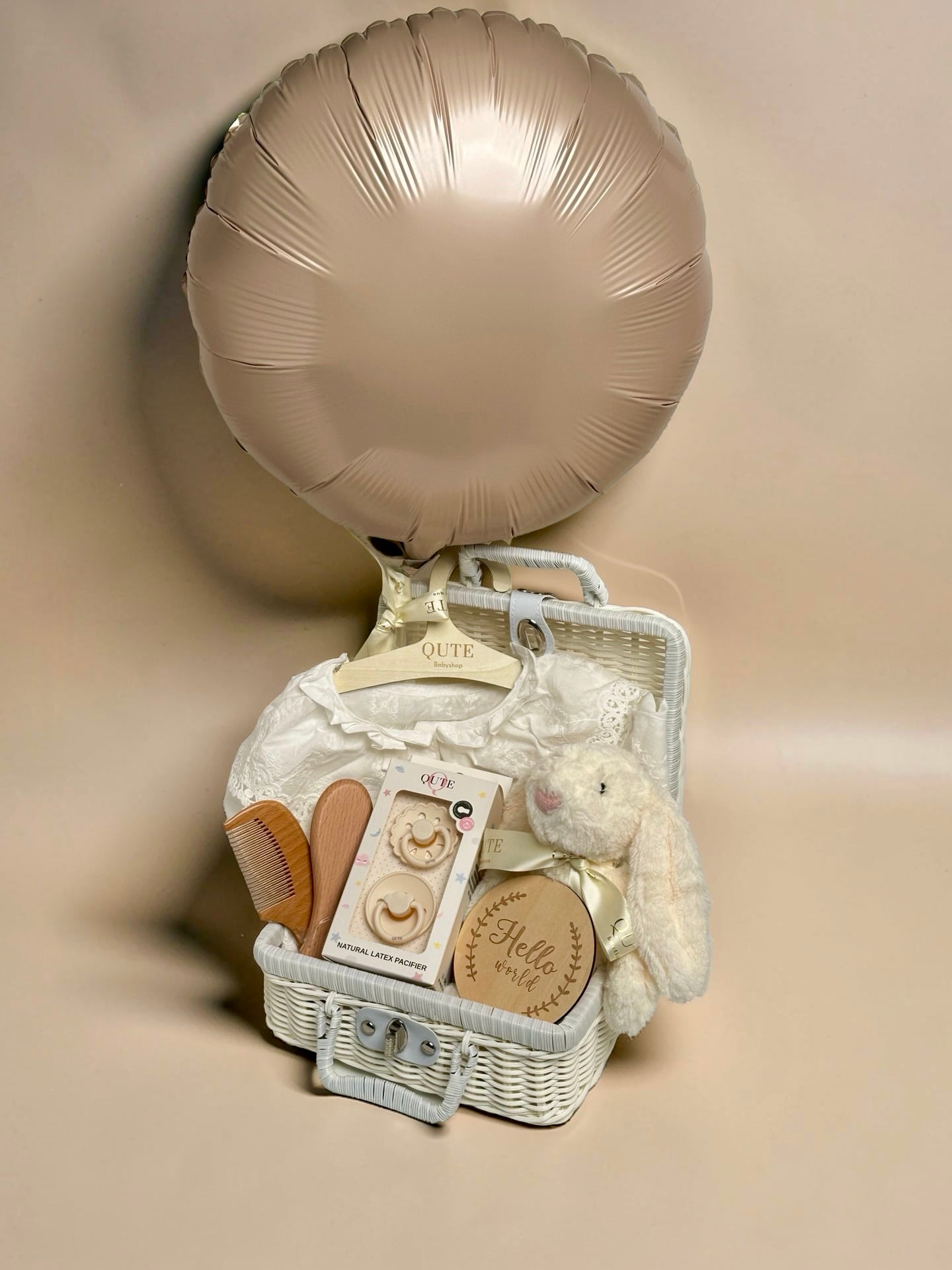 Deluxe white baby girl gift set in white woven suitcase hamper featuring champagne gold foil balloon, ivory lace collar dress, plush bunny toy, Qute natural latex pacifiers, wooden hairbrush comb set, and milestone disc by Qute Gifts Doha Qatar