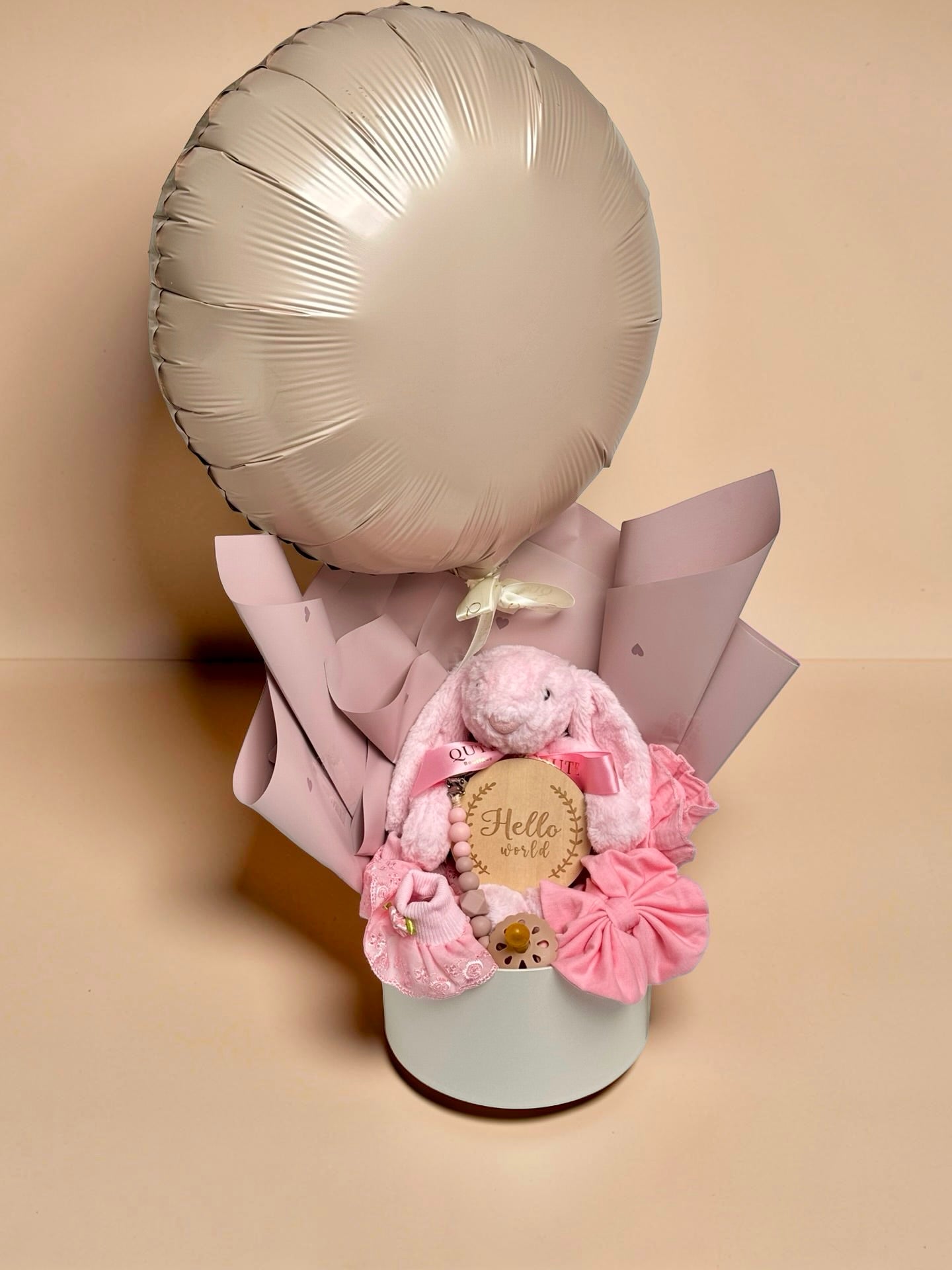 Blush pink baby girl gift bouquet box featuring champagne gold foil balloon, plush pink bunny toy, wooden milestone disc, pink baby booties, headband, and pacifier clip by Qute Gifts Doha Qatar