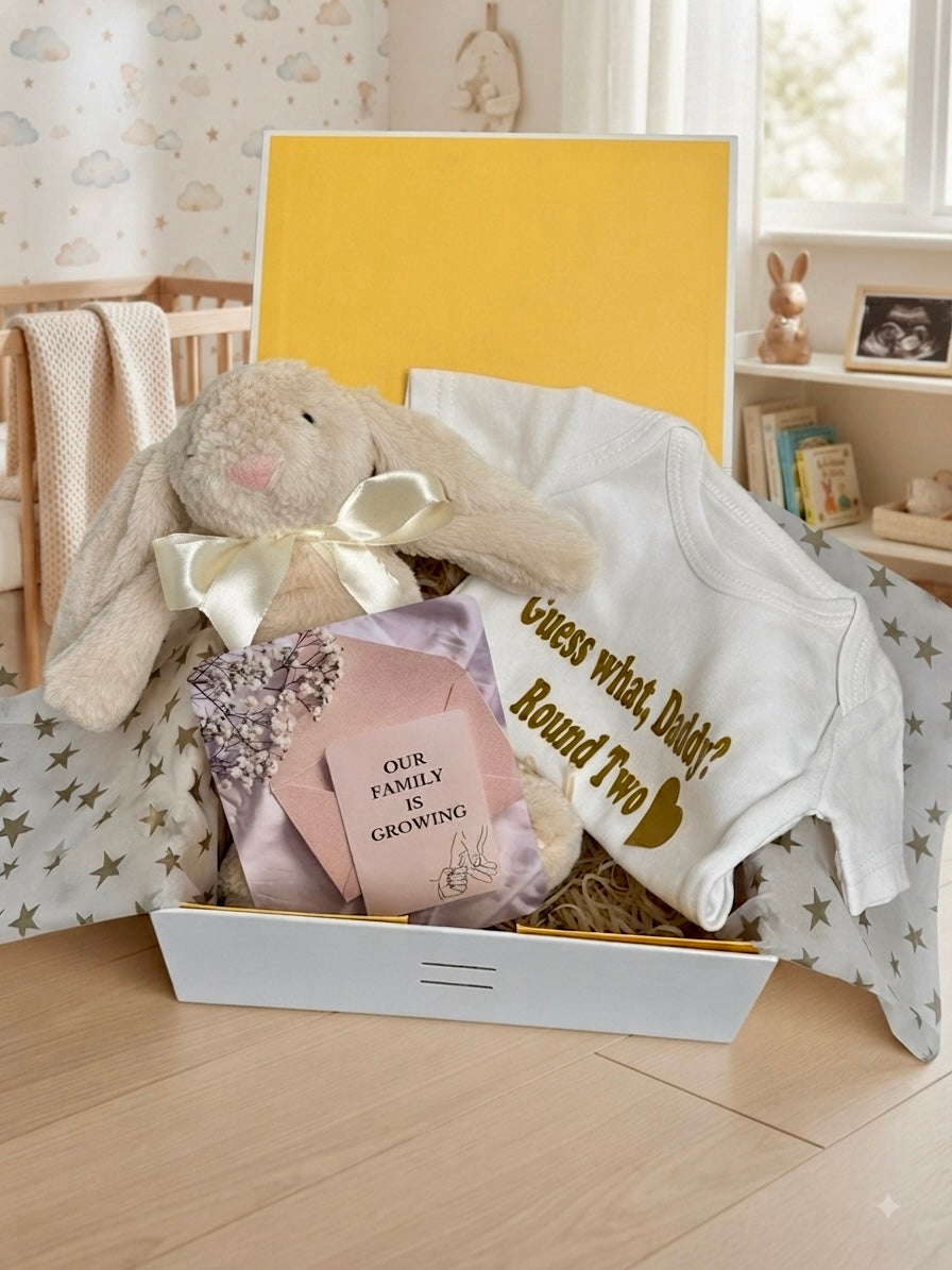 Pregnancy announcement gift box for husband featuring white onesie printed with Guess What Daddy Round Two, plush bunny toy, and Our Family Is Growing reveal card by Qute Gifts Doha Qatar