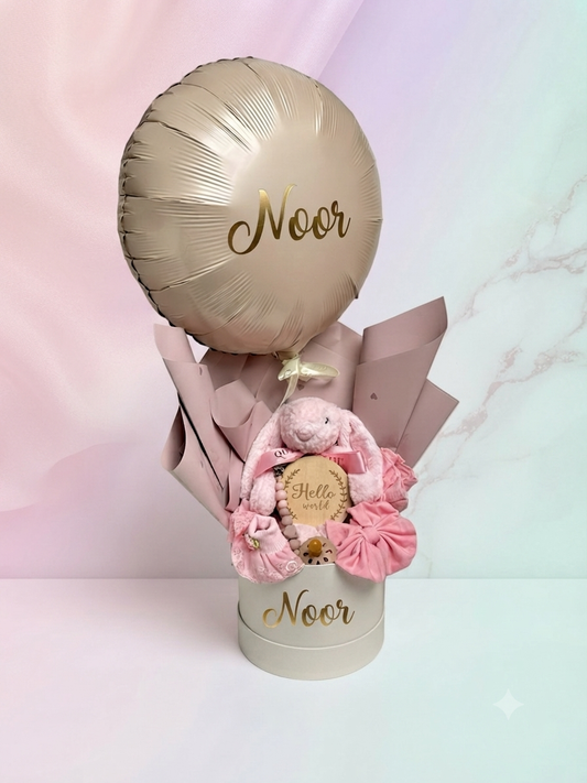 A personalized baby shower gift basket set featuring a metallic gold foil balloon, a pink plush teddy bear, pink satin bows, and a round gift box with the name 'Noor' printed on it, set against a pastel pink and mint background.