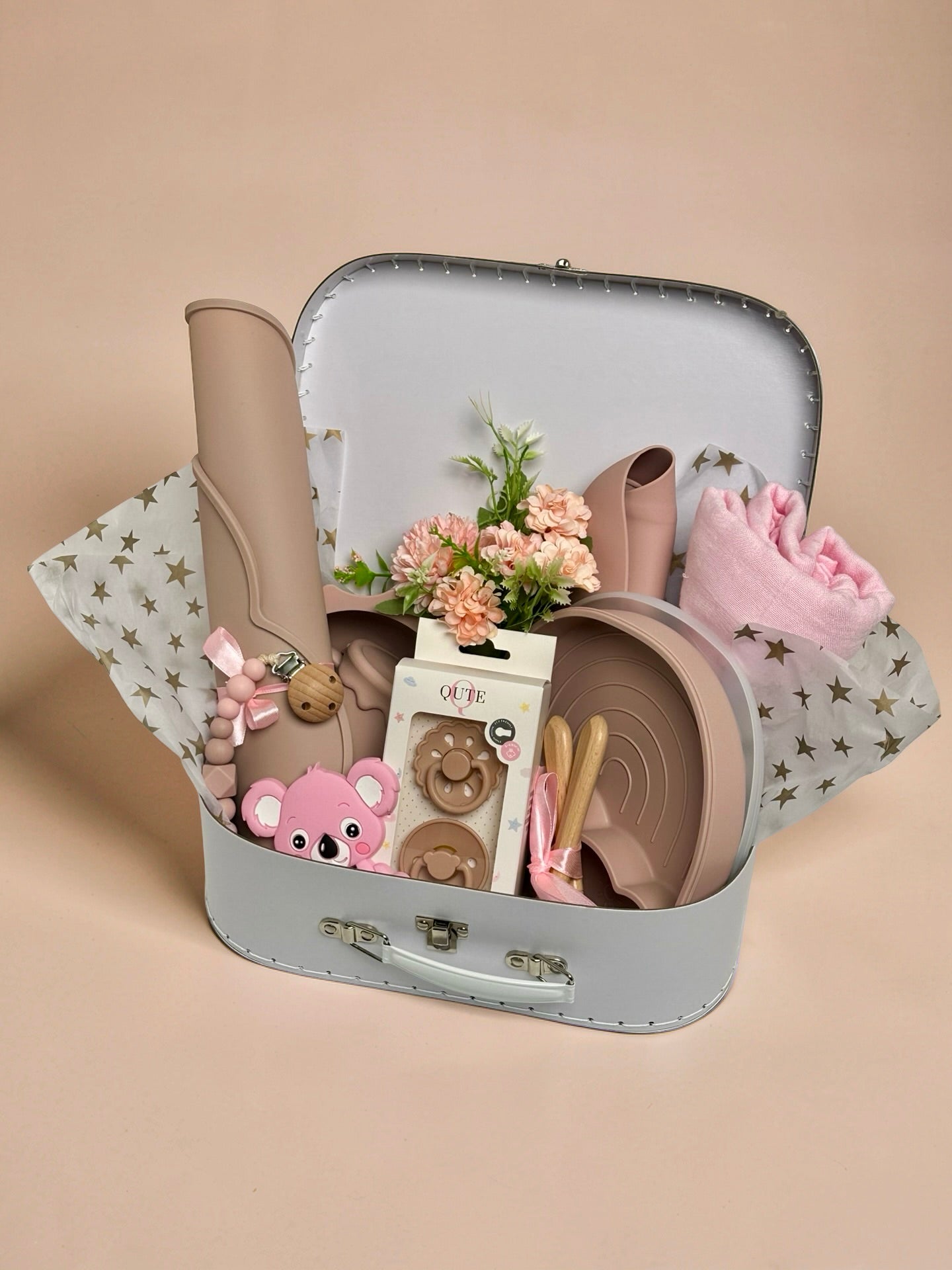 Luxury baby girl newborn and toddler gift set inside a white keepsake suitcase featuring a pink organic cotton swaddle, divided rainbow silicone weaning plate, sun bowl, placemat, training cup, bib, natural latex pacifiers, pink koala teether, and floral arrangement in Doha Qatar