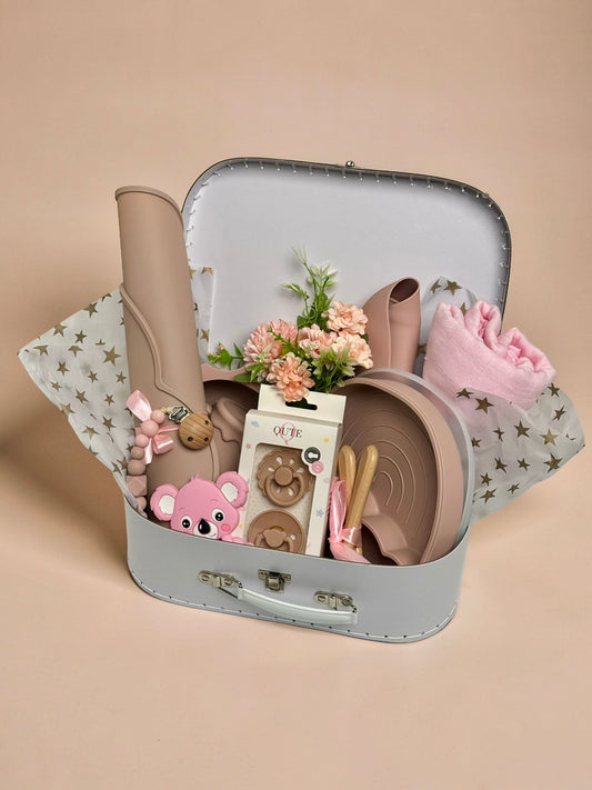 Luxury baby girl newborn and toddler gift set inside a white keepsake suitcase featuring a pink organic cotton swaddle, divided rainbow silicone weaning plate, sun bowl, placemat, training cup, bib, natural latex pacifiers, pink koala teether, and floral arrangement in Doha Qatar