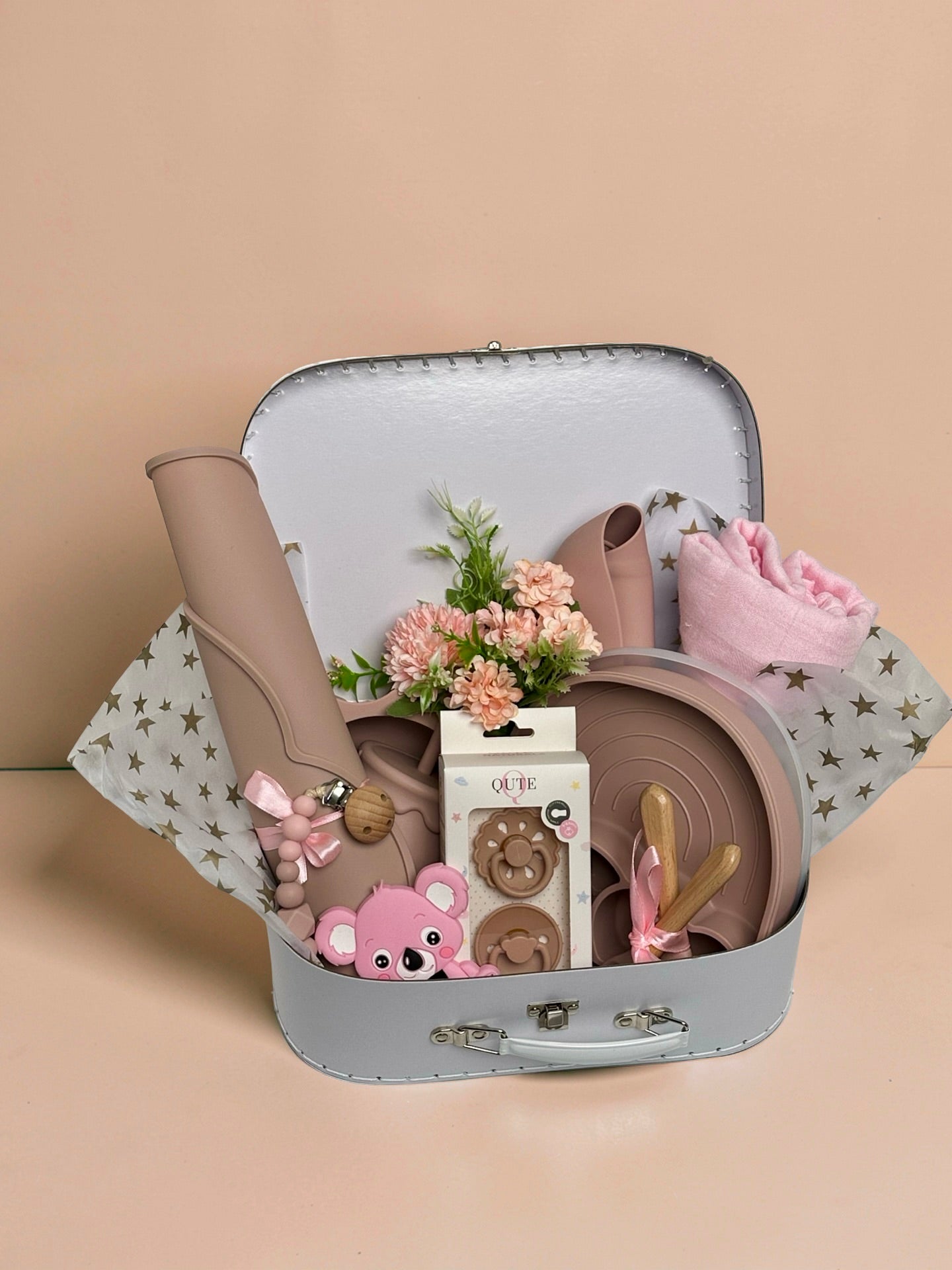 Luxury baby girl newborn and toddler gift set inside a white keepsake suitcase featuring a pink organic cotton swaddle, divided rainbow silicone weaning plate, sun bowl, placemat, training cup, bib, natural latex pacifiers, pink koala teether, and floral arrangement in Doha Qatar