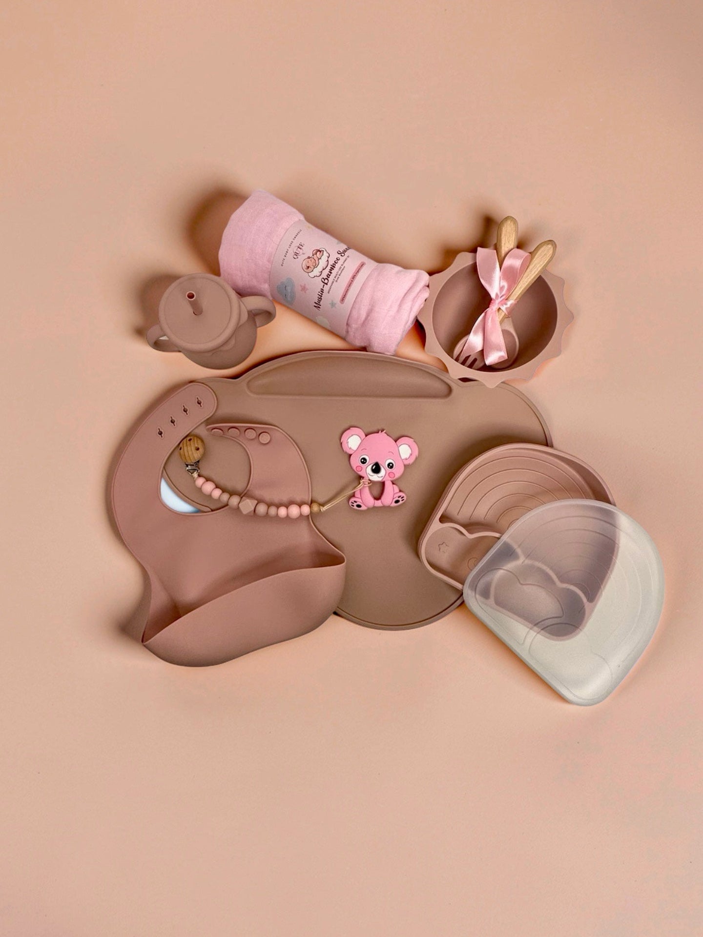 Baby girl silicone weaning and feeding set flat lay in dusty pink, featuring a large placemat, divided rainbow plate with clear lid, sun-shaped bowl with wooden spoon and fork tied with a pink ribbon, two-handled training straw cup, silicone bib, boxed natural latex pacifiers, pink koala teether with beaded pacifier clip, and a rolled pink muslin swaddle blanket in Doha Qatar