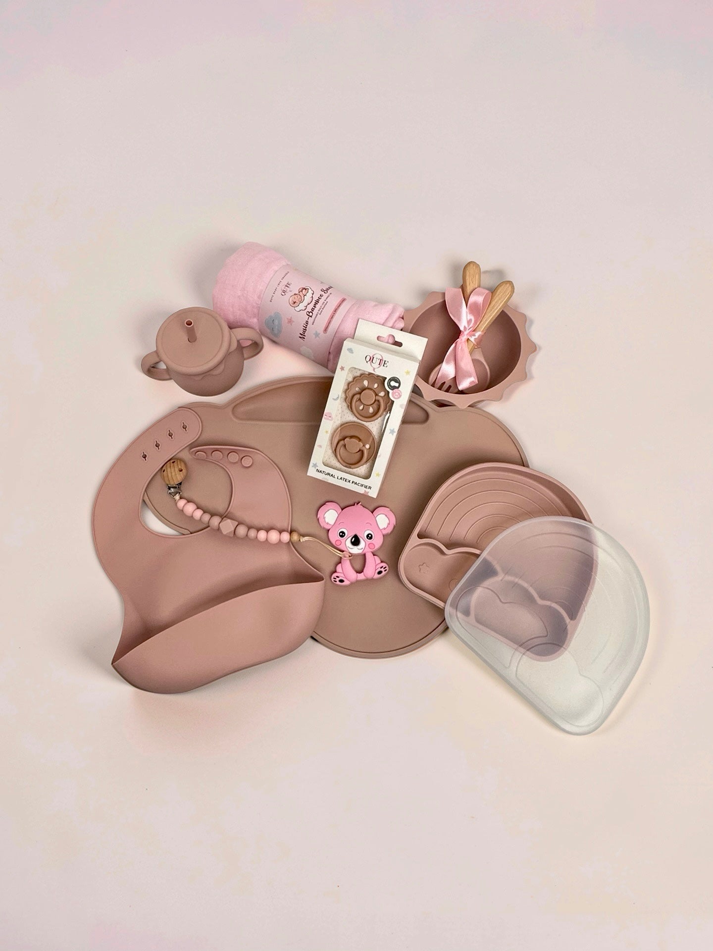Baby girl silicone weaning and feeding set flat lay in dusty pink, featuring a large placemat, divided rainbow plate with clear lid, sun-shaped bowl with wooden spoon and fork tied with a pink ribbon, two-handled training straw cup, silicone bib, boxed natural latex pacifiers, pink koala teether with beaded pacifier clip, and a rolled pink muslin swaddle blanket in Doha Qatar