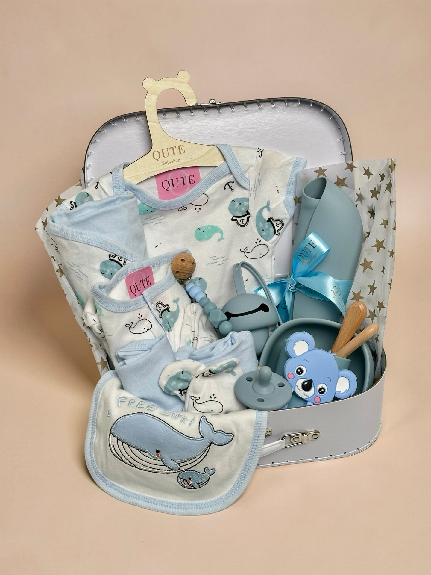 Luxury baby boy gift set inside a white keepsake suitcase featuring a whale-themed clothing outfit on a branded wooden hanger, matching whale bib, blue silicone feeding bowl, wooden spoon set, blue koala teether, pacifier, beaded clip, and silicone bib in Doha Qatar