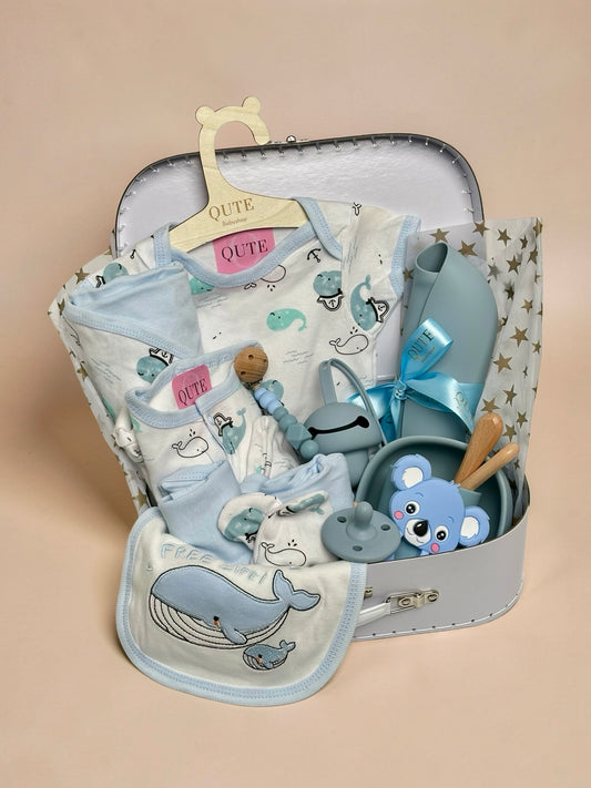 Luxury baby boy gift set inside a white keepsake suitcase featuring a whale-themed clothing outfit on a branded wooden hanger, matching whale bib, blue silicone feeding bowl, wooden spoon set, blue koala teether, pacifier, beaded clip, and silicone bib in Doha Qatar