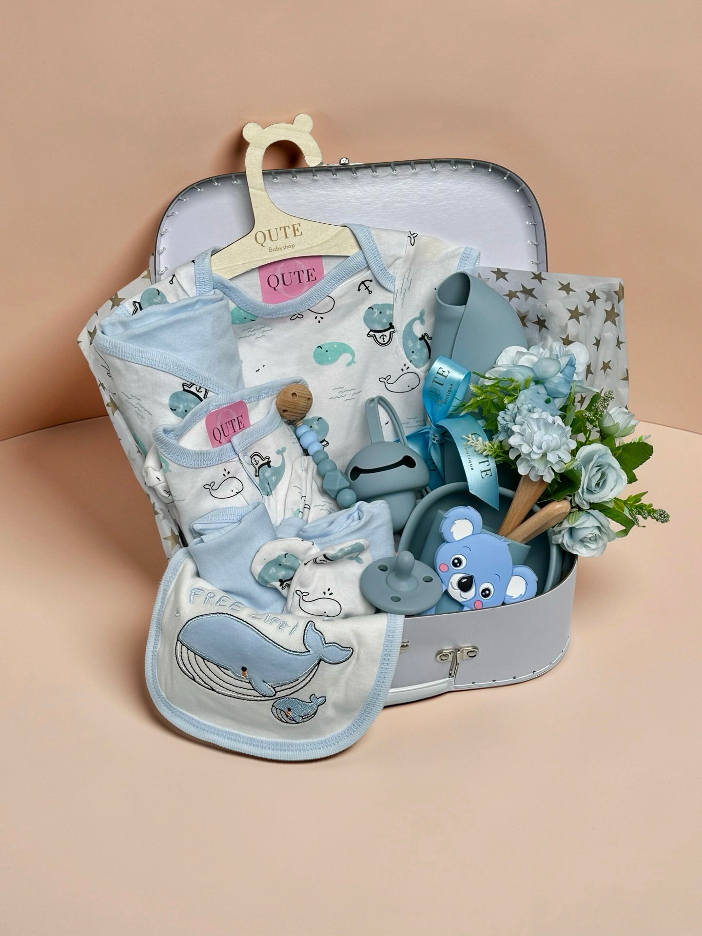 Luxury baby boy gift set inside a white keepsake suitcase featuring a whale-themed clothing outfit on a branded wooden hanger, matching whale bib, blue silicone feeding bowl, wooden spoon set, blue koala teether, pacifier, beaded clip, and silicone bib in Doha Qatar