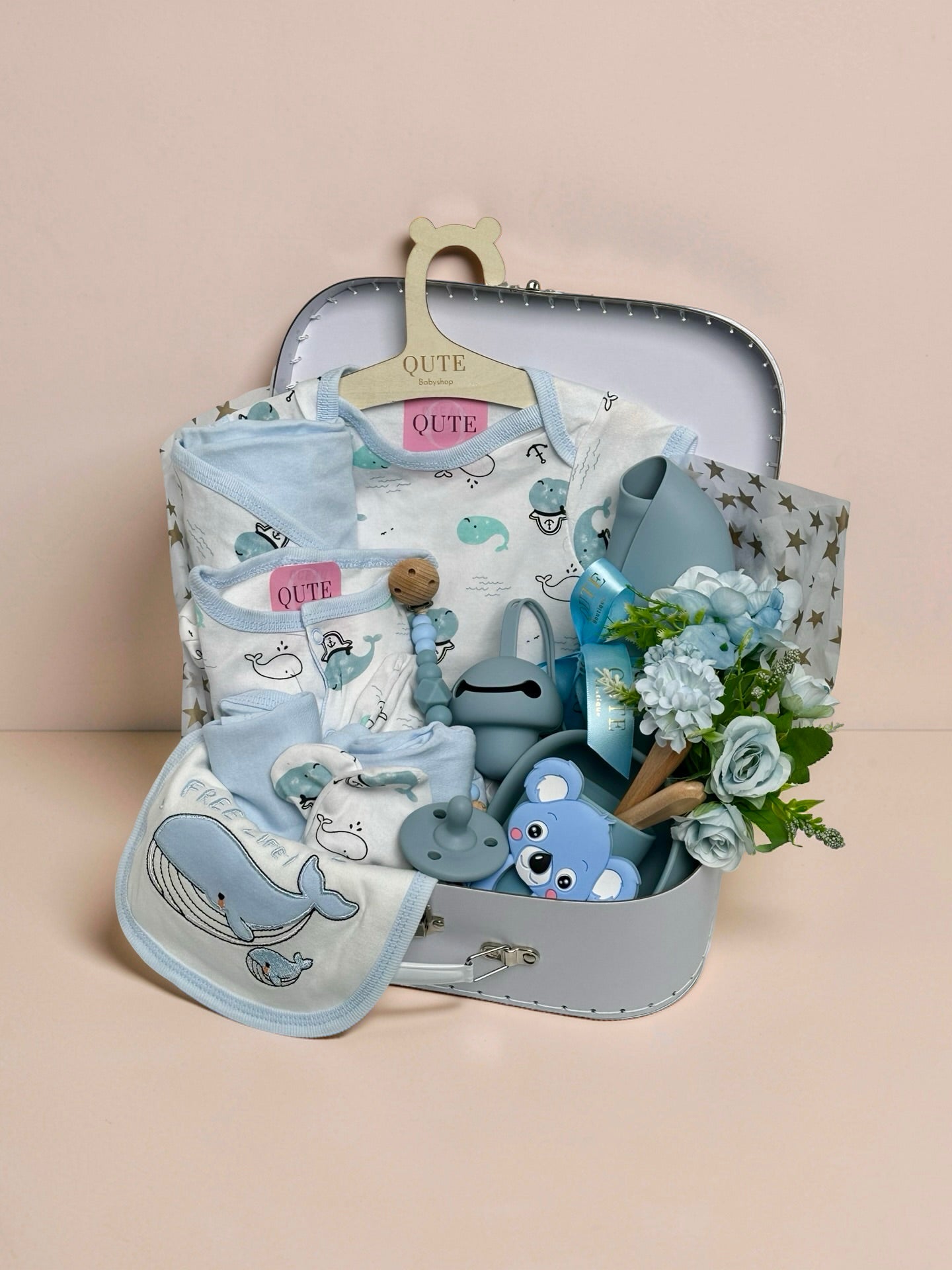 Luxury baby boy gift set inside a white keepsake suitcase featuring a whale-themed clothing outfit on a branded wooden hanger, matching whale bib, blue silicone feeding bowl, wooden spoon set, blue koala teether, pacifier, beaded clip, and silicone bib in Doha Qatar