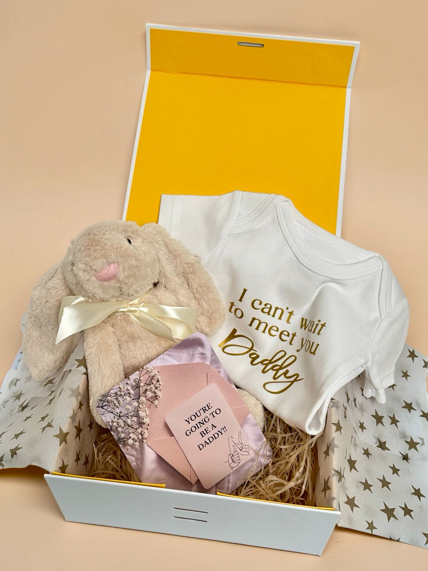 Pregnancy announcement gift box featuring a white baby bodysuit with gold foil text I can't wait to meet you Daddy, a soft cream plush bunny with satin ribbon, a pink envelope with a You're Going to Be a Daddy announcement card, and star-patterned tissue paper in a luxury gift box in Doha Qatar