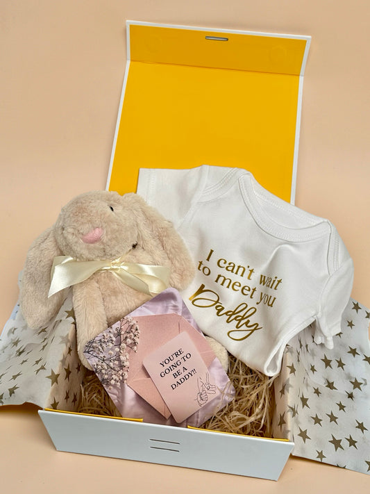 Pregnancy announcement gift box featuring a white baby bodysuit with gold foil text I can't wait to meet you Daddy, a soft cream plush bunny with satin ribbon, a pink envelope with a You're Going to Be a Daddy announcement card, and star-patterned tissue paper in a luxury gift box in Doha Qatar