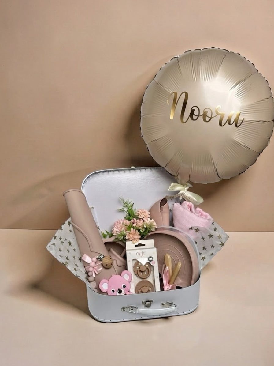 Personalised baby girl gift set in a white keepsake suitcase featuring a metallic gold custom balloon with the name Noora, soft pink swaddle blanket, silicone tableware set, pink koala teether, pacifier set, and small floral arrangement in Doha Qatar