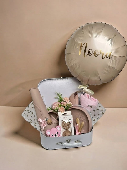 Personalised baby girl gift set in a white keepsake suitcase featuring a metallic gold custom balloon with the name Noora, soft pink swaddle blanket, silicone tableware set, pink koala teether, pacifier set, and small floral arrangement in Doha Qatar