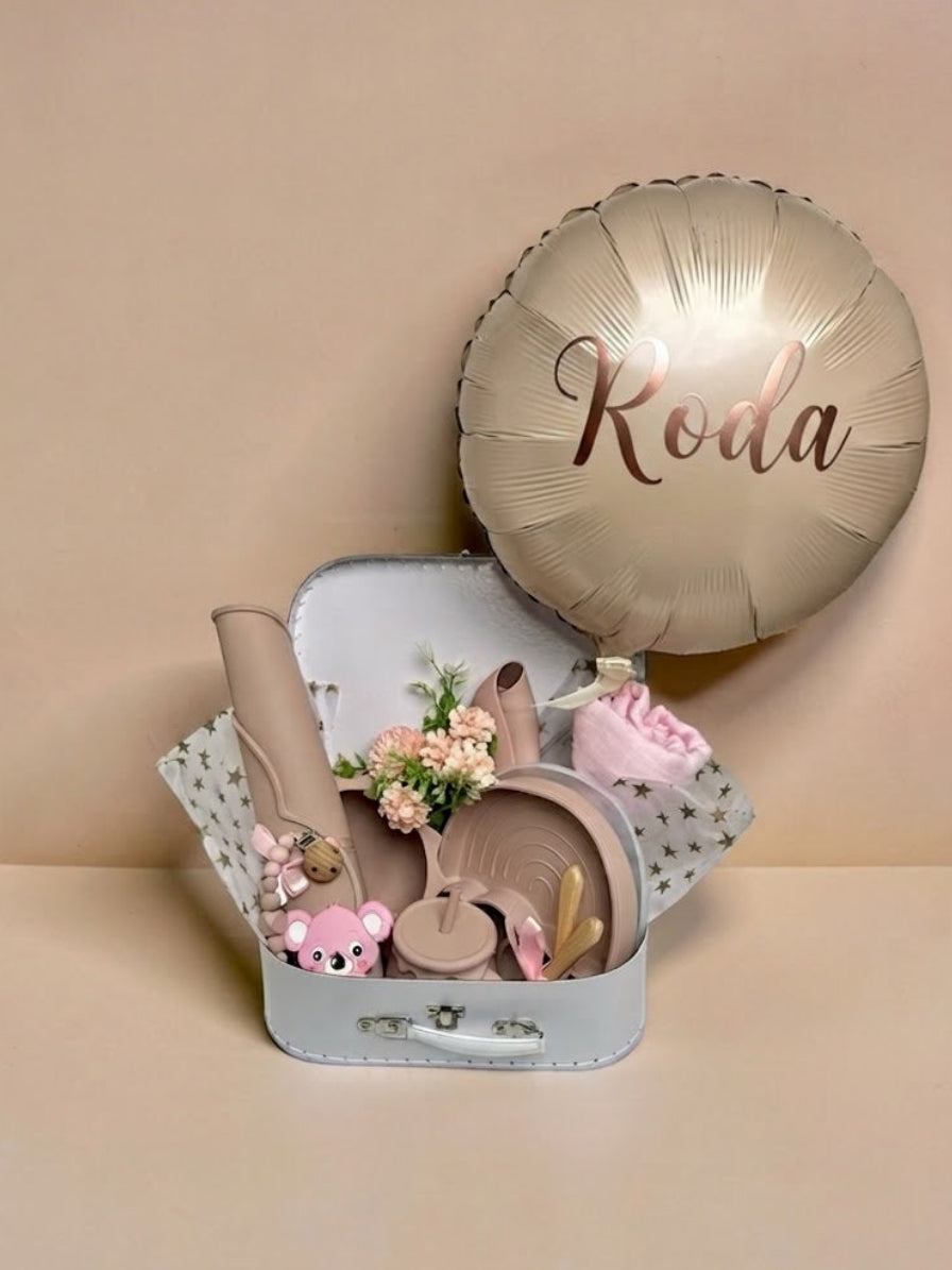 Personalised baby girl gift set in a white keepsake suitcase featuring a metallic champagne-gold balloon with the name Roda printed on it, silicone weaning tableware, pink koala teether, swaddle, and floral arrangement in Doha Qatar