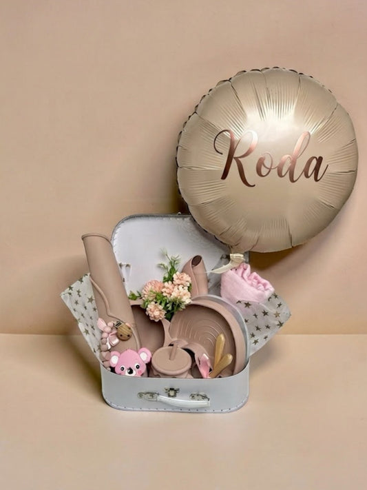 Personalised baby girl gift set in a white keepsake suitcase featuring a metallic champagne-gold balloon with the name Roda printed on it, silicone weaning tableware, pink koala teether, swaddle, and floral arrangement in Doha Qatar