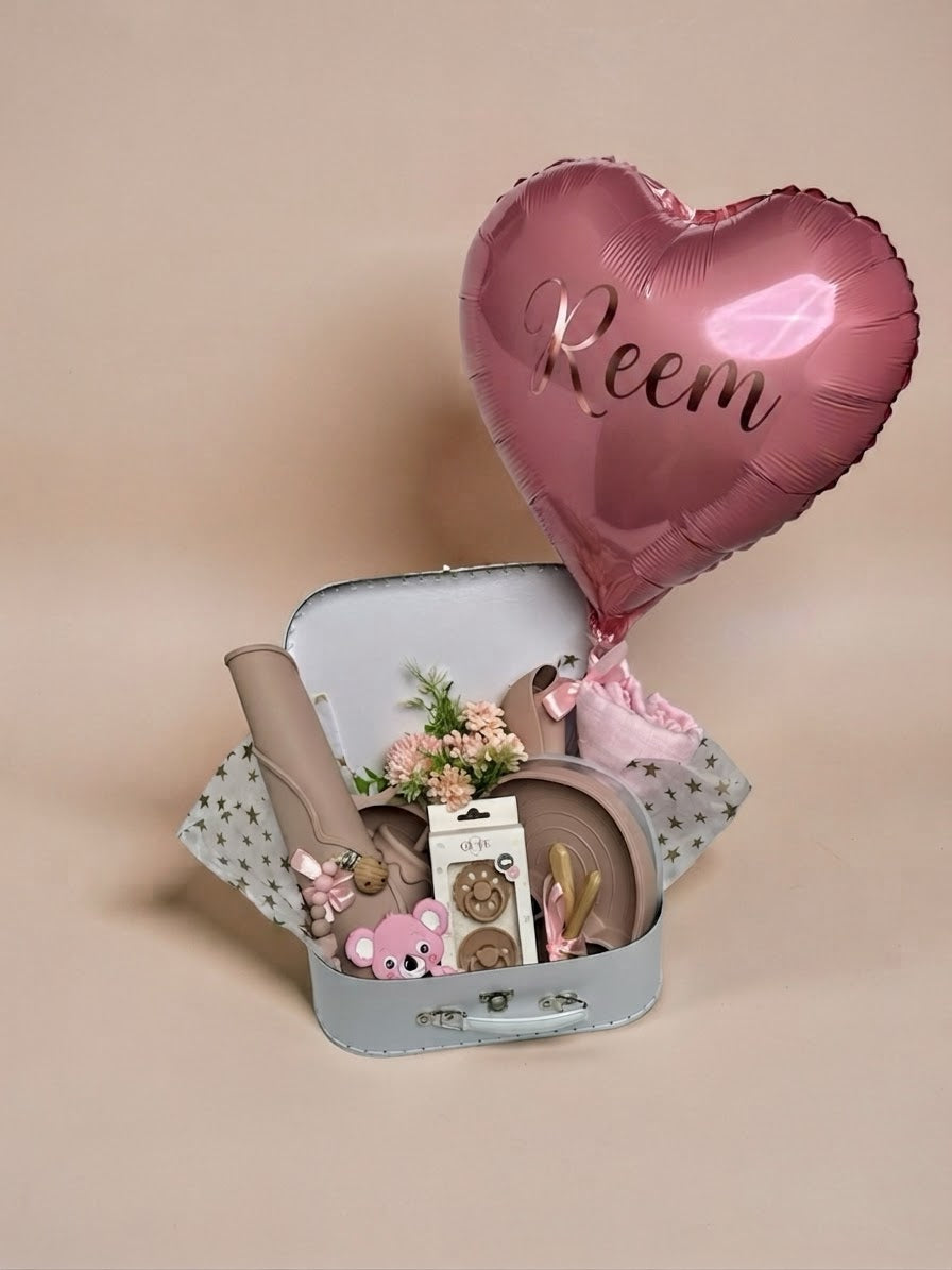 Personalised baby girl gift set in a white keepsake suitcase featuring a metallic pink heart balloon with the name Reem printed on it, pink silicone weaning tableware, boxed natural latex pacifiers, pink koala teether, pink swaddle, and small floral arrangement in Doha Qatar
