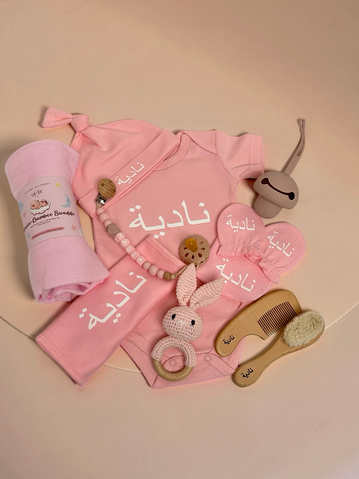 Personalised baby girl clothing and care set flat lay in pink, featuring a custom baby bodysuit, matching hat, mittens, bib, and a rolled muslin-bamboo swaddle blanket all embroidered with the name Nadiya in Arabic script, alongside a beaded pacifier clip spelling Nadiya in English, a pacifier, a pink crochet bunny rattle, a silicone dummy, and a wooden hairbrush and comb set engraved with Arabic text in Doha Qatar