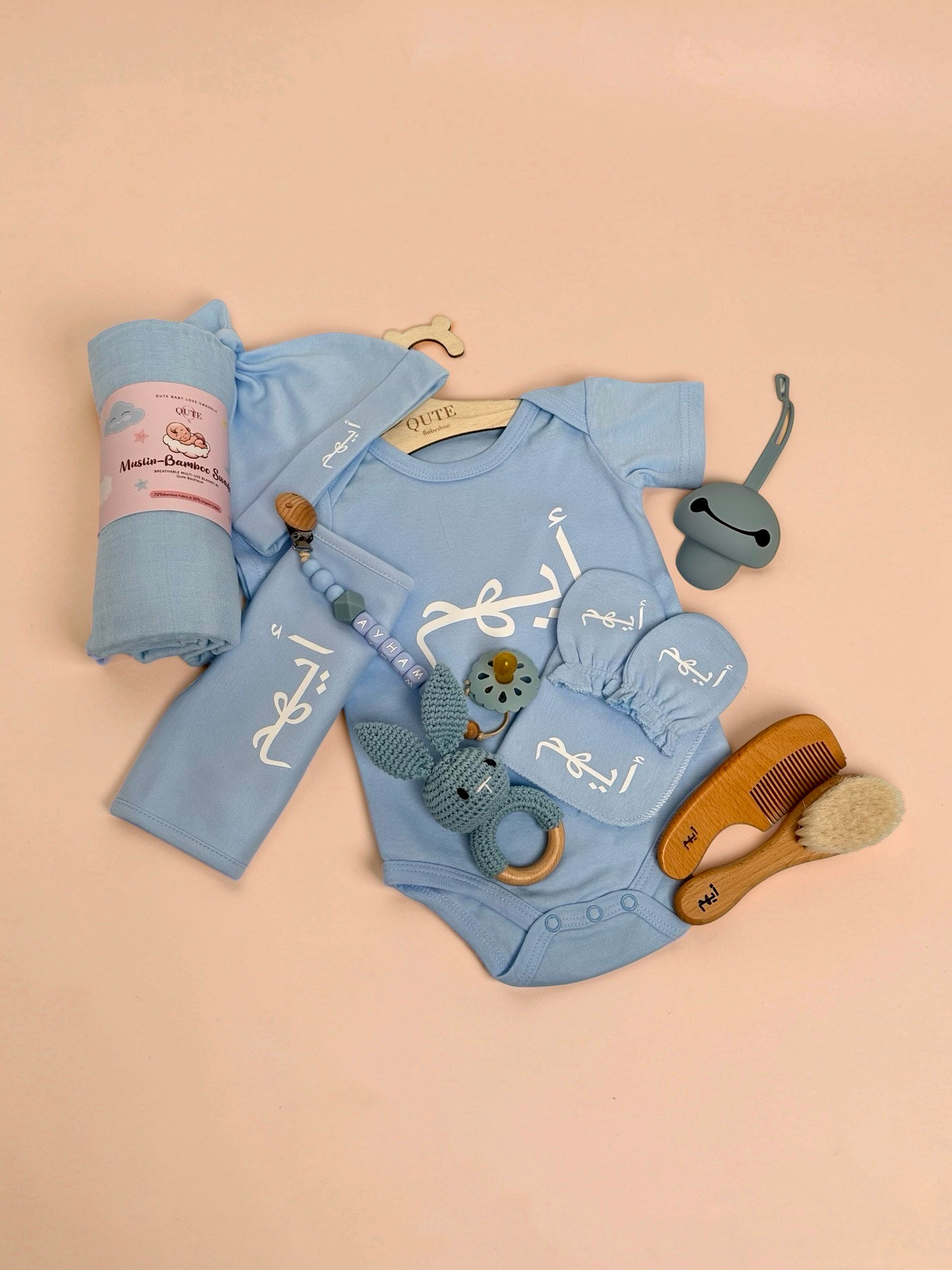 Personalised baby boy clothing and care set flat lay in blue, featuring a custom baby bodysuit, matching hat, mittens, bib, and a rolled muslin-bamboo swaddle blanket all embroidered with the name Ayham in Arabic script, alongside a beaded pacifier clip spelling Ayham in English, a pacifier, a blue crochet bunny rattle, a grey silicone dummy, and a wooden hairbrush and comb set engraved with Arabic text in Doha Qatar