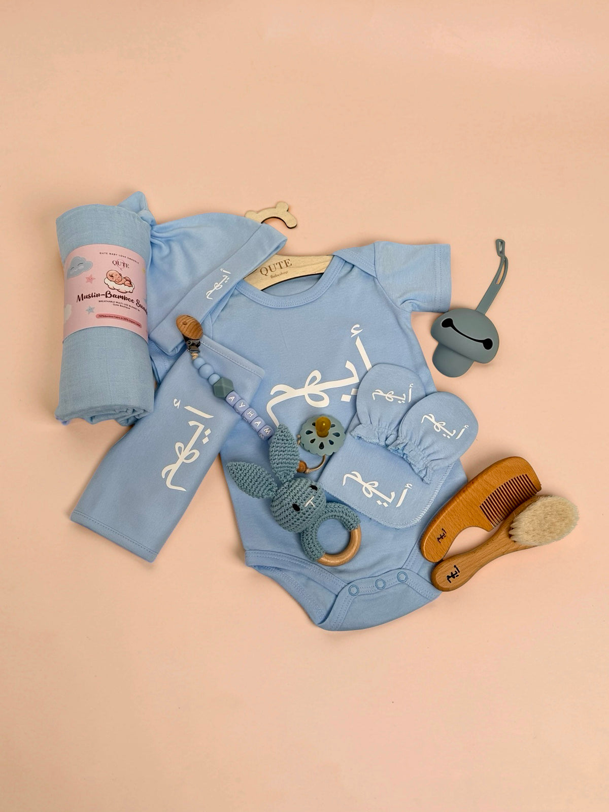 Personalised baby boy clothing and care set flat lay in blue, featuring a custom baby bodysuit, matching hat, mittens, bib, and a rolled muslin-bamboo swaddle blanket all embroidered with the name Ayham in Arabic script, alongside a beaded pacifier clip spelling Ayham in English, a pacifier, a blue crochet bunny rattle, a grey silicone dummy, and a wooden hairbrush and comb set engraved with Arabic text in Doha Qatar
