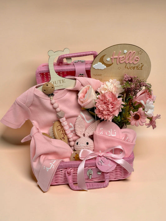 Personalised baby girl gift basket in a pink wicker suitcase featuring a pink bodysuit with a beaded pacifier clip reading the name Nadiya, a wooden baby brush with Arabic text, a small crochet bunny toy, pink floral arrangement, a Hello World wooden milestone plaque, and a pink ribbon tag in Doha Qatar