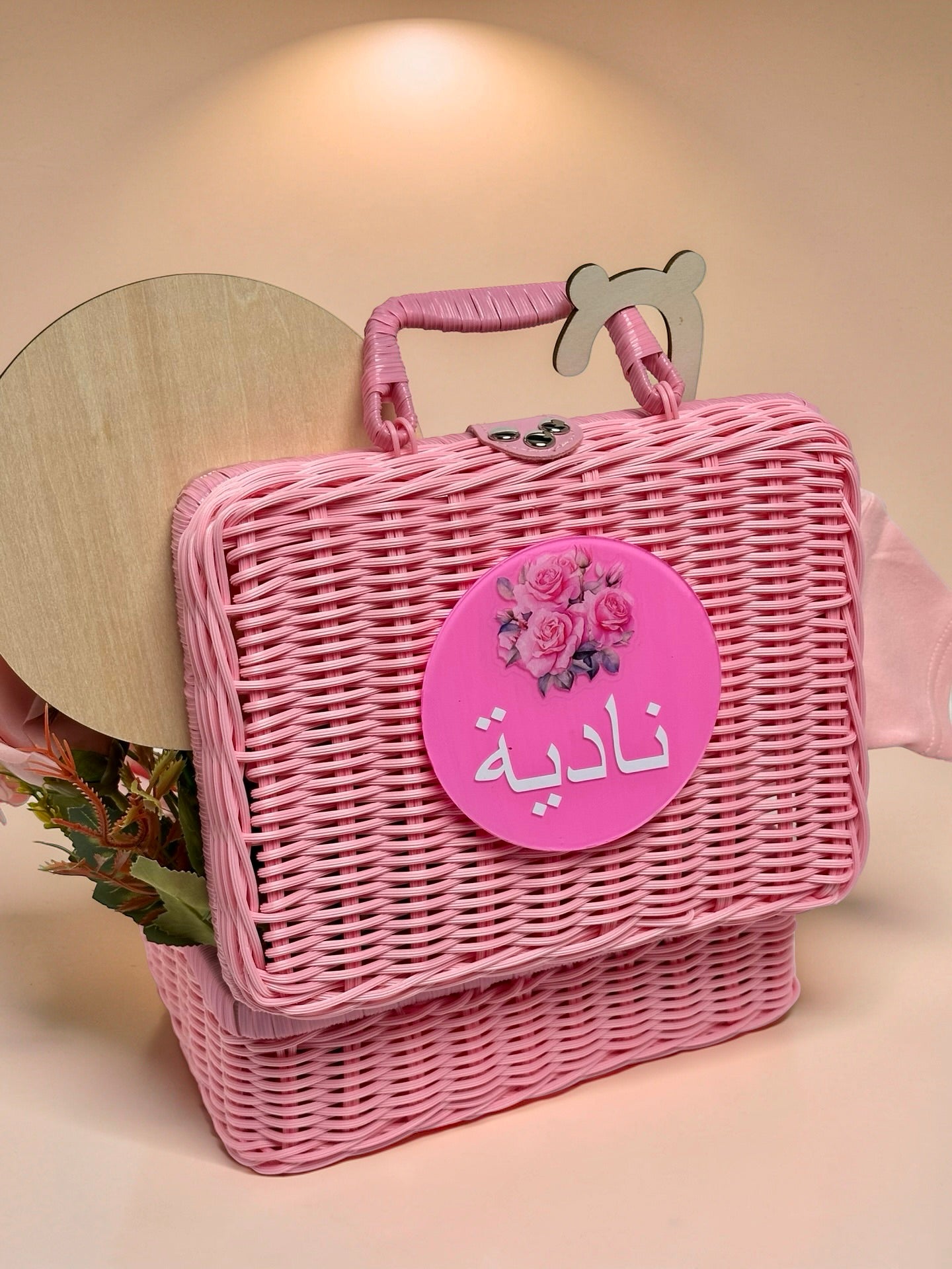 Personalised baby girl pink wicker basket and suitcase featuring a round pink plaque on the front with an illustration of roses and the name Nadiya written in Arabic script, with a round wooden milestone disc and floral arrangement peeking out from behind in Doha Qatar