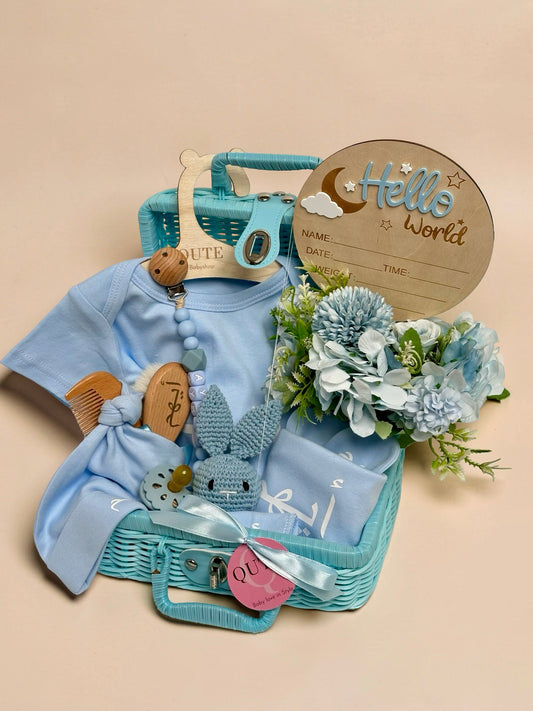 Personalised baby boy gift basket in a blue wicker suitcase featuring a light blue bodysuit with a blue beaded pacifier clip spelling the name Ayham, a wooden baby brush engraved with Arabic text, a small blue crochet bunny toy, blue floral arrangement, a Hello World wooden milestone plaque, and a blue ribbon tag in Doha Qatar