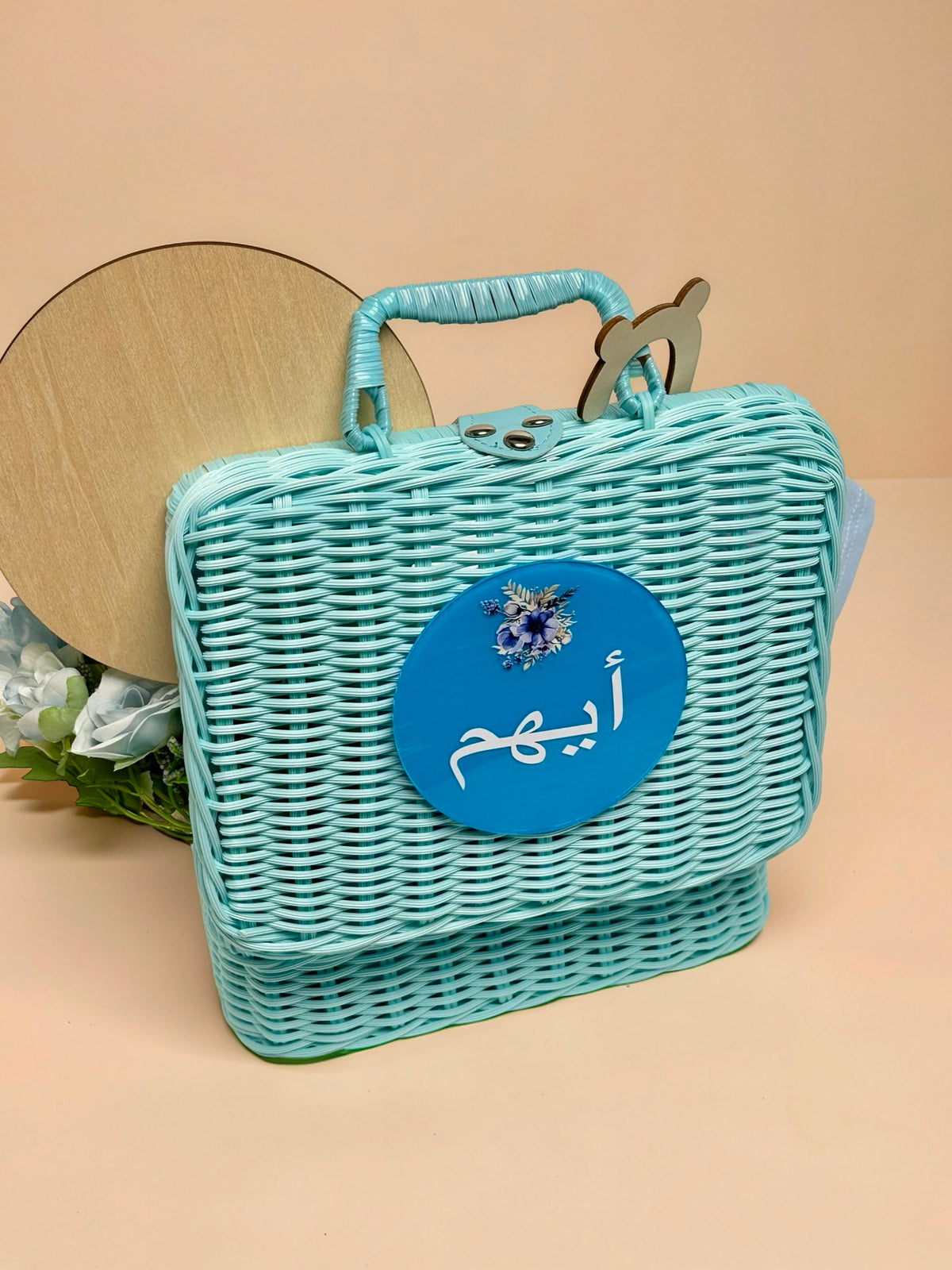 Personalised baby boy blue wicker basket and suitcase featuring a round blue plaque on the front with an illustration of blue flowers and the name Ayham written in Arabic script, with a round wooden milestone disc and floral arrangement peeking out from behind in Doha Qatar