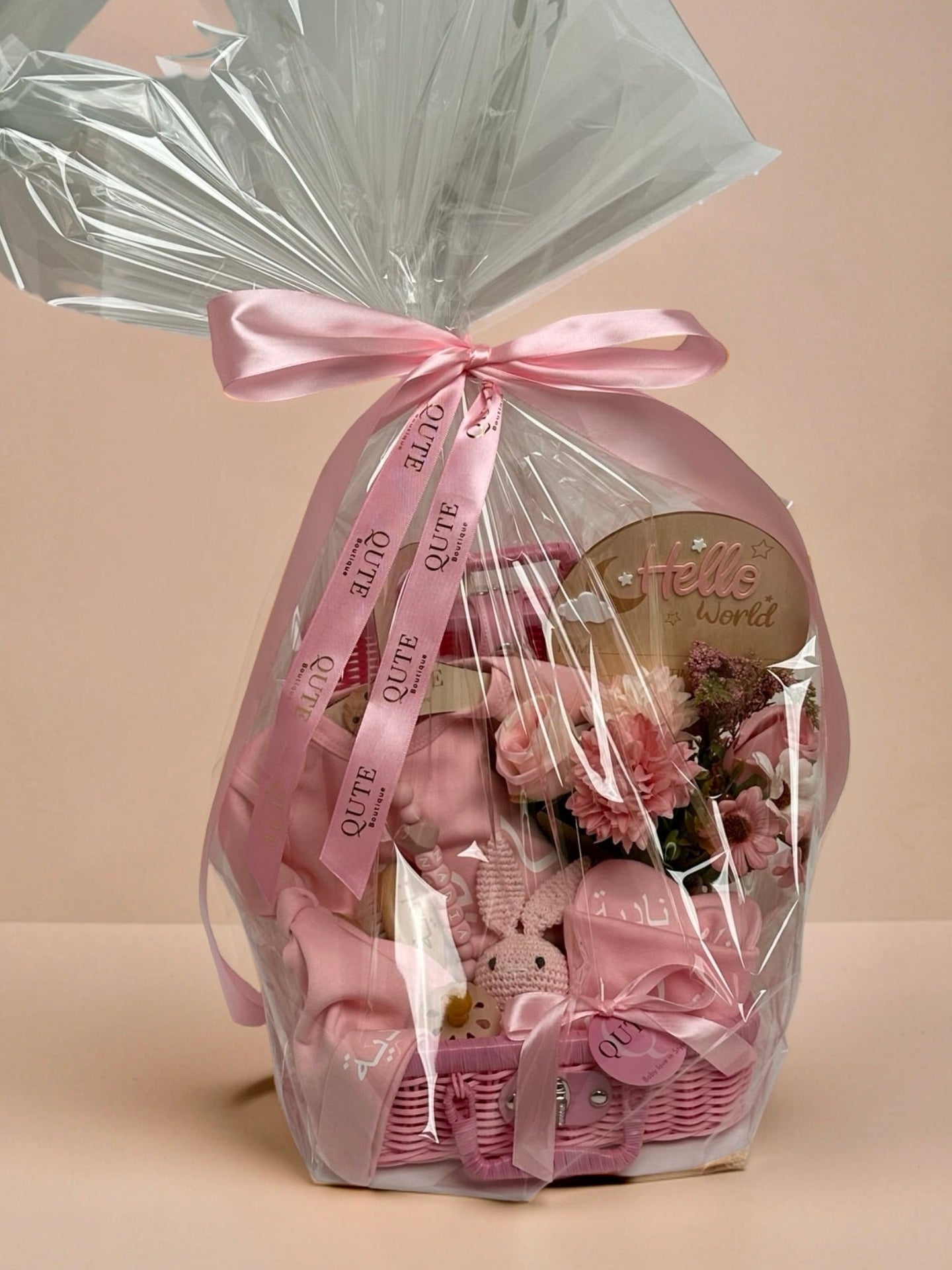 Personalised baby girl gift basket wrapped in clear cellophane and tied with a custom pink ribbon printed with the Qute Boutique logo, showcasing a pink wicker suitcase filled with baby essentials, a crochet bunny toy, a Hello World wooden plaque, and floral arrangements in Doha Qatar