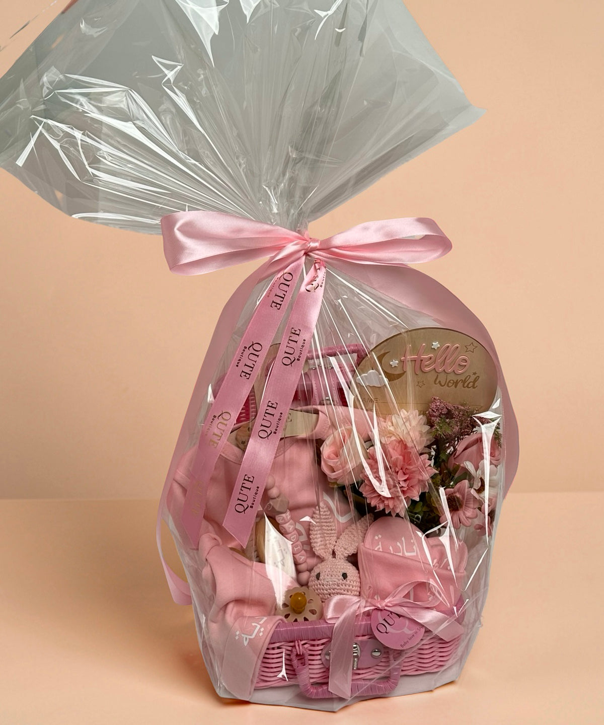Personalised baby girl gift basket wrapped in clear cellophane and tied with a custom pink ribbon printed with the Qute Boutique logo, showcasing a pink wicker suitcase filled with baby essentials, a crochet bunny toy, a Hello World wooden plaque, and floral arrangements in Doha Qatar