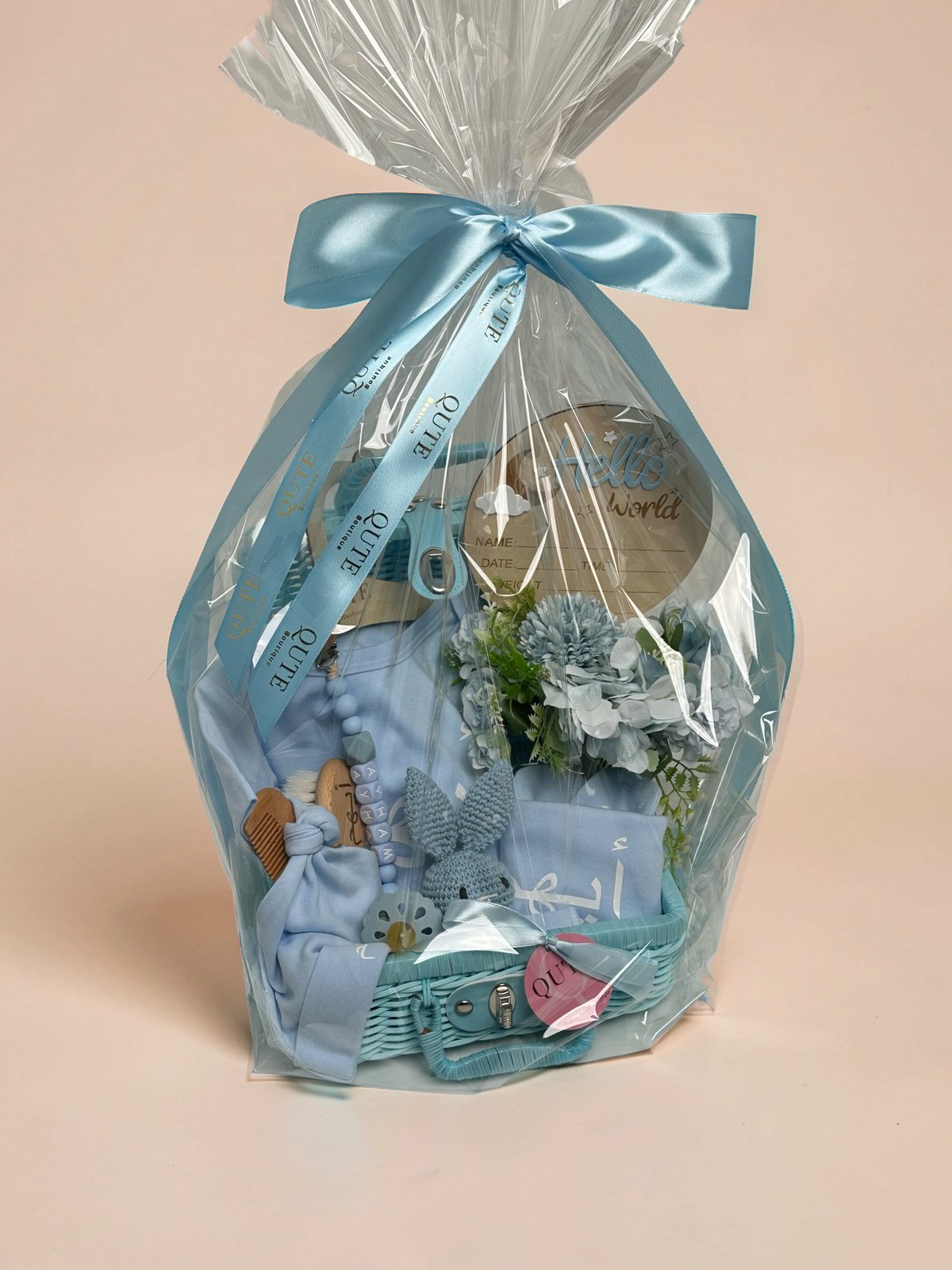Personalised baby boy gift basket wrapped in clear cellophane and tied with a custom blue satin ribbon printed with the Qute Gifts logo, showcasing a blue wicker suitcase filled with baby essentials, a crochet bunny toy, a Hello World wooden plaque, and blue floral arrangements in Doha Qatar