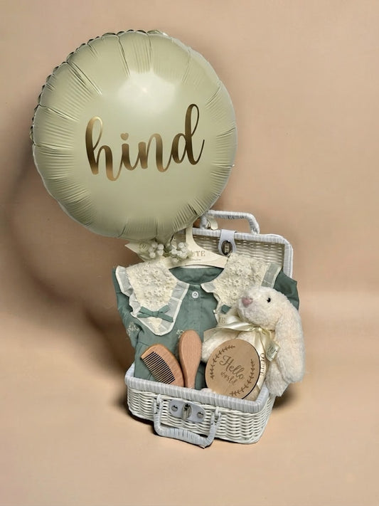 Personalised baby gift set in a white wicker suitcase featuring a sage green round foil balloon printed with the name Hind in gold lettering, a sage green dress with white lace collar on a Qute hanger, a plush white bunny toy, a wooden Hello World milestone disc, and a wooden hairbrush and comb set in Doha Qatar