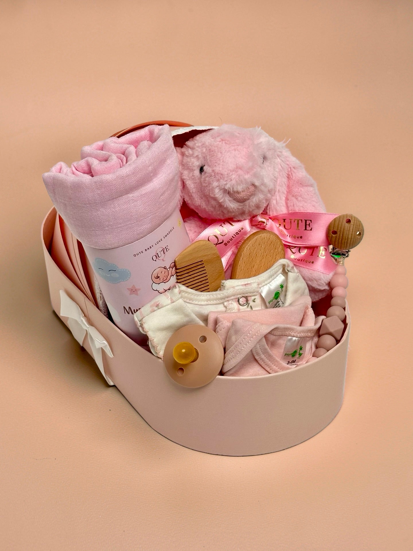 Luxury baby girl newborn gift hamper in a cradle-shaped pink box by Qute Qatar, featuring a pink swaddle, plush bunny, baby clothes, and wooden accessories for delivery in Doha.