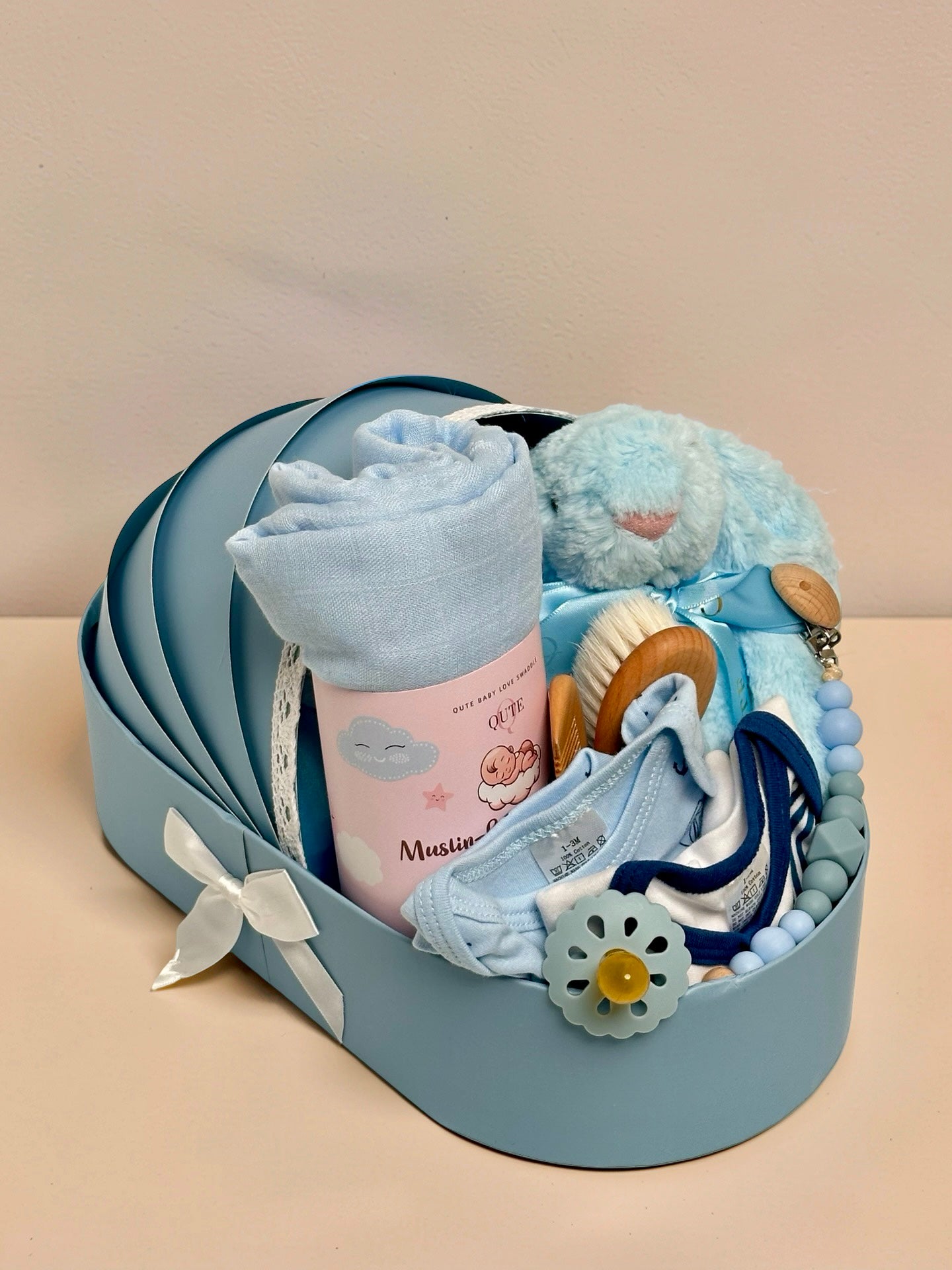 Luxury baby boy newborn gift hamper in a cradle-shaped blue box by Qute Qatar, featuring a blue swaddle, plush bunny, baby clothes, and wooden accessories for delivery in Doha.
