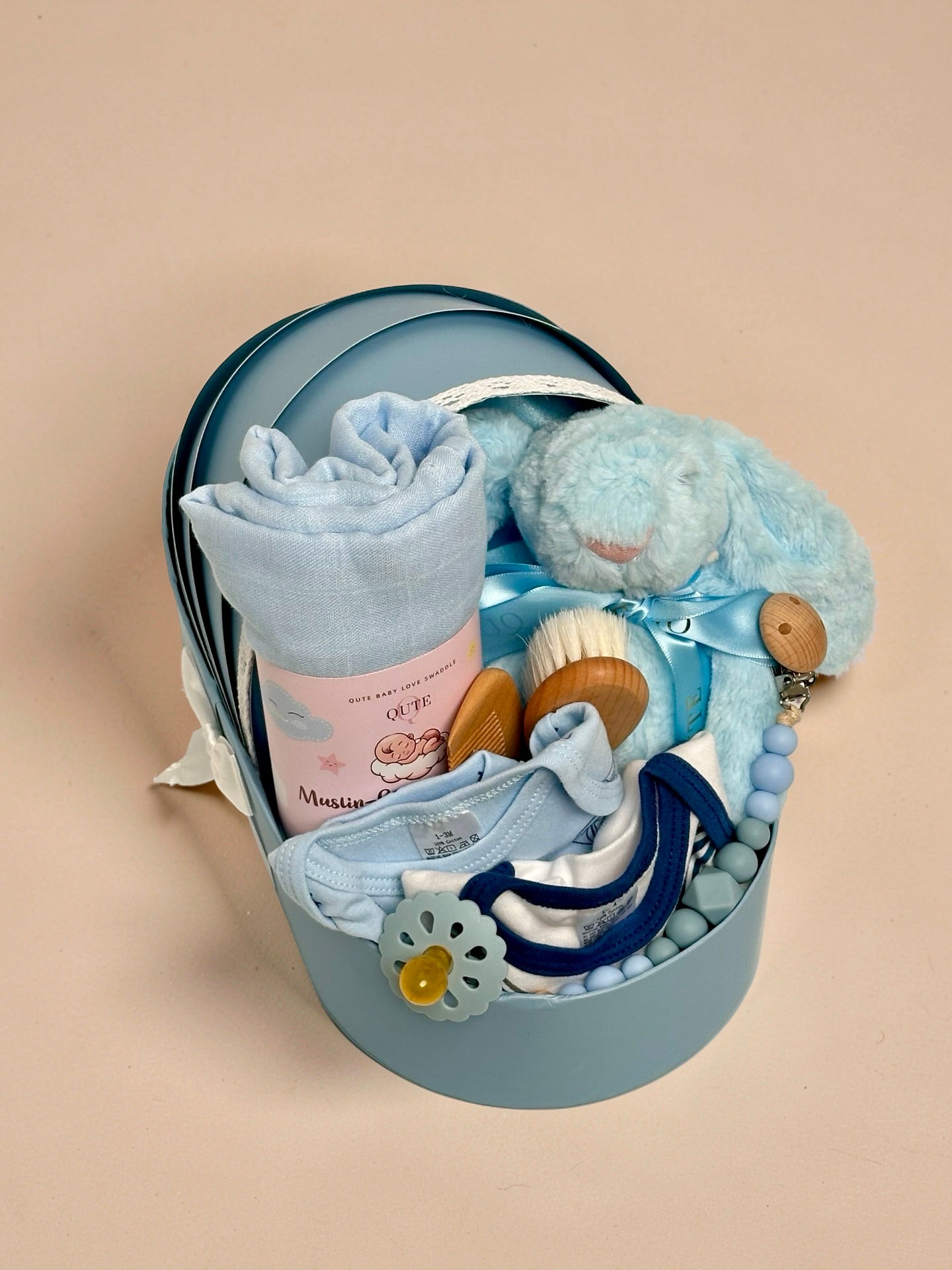 Luxury baby boy newborn gift hamper in a cradle-shaped blue box by Qute Qatar, featuring a blue swaddle, plush bunny, baby clothes, and wooden accessories for delivery in Doha.