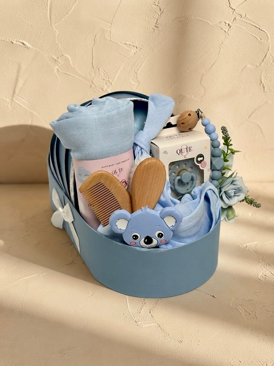 Luxury baby boy newborn gift hamper in a sky blue cradle-shaped box by Qute Qatar, featuring a blue swaddle, koala teether, baby clothes, and wooden accessories for delivery in Doha.