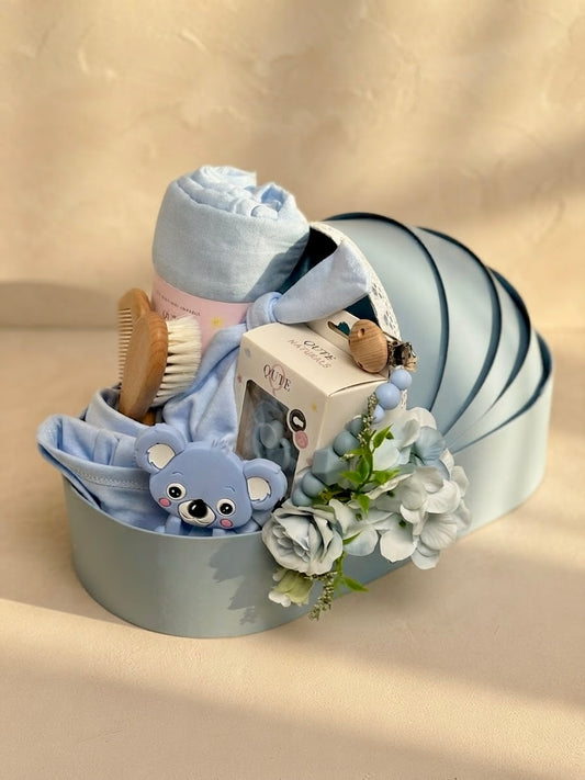 Luxury baby boy newborn gift hamper in a sky blue cradle-shaped box by Qute Qatar, featuring a blue swaddle, koala teether, baby clothes, and wooden accessories for delivery in Doha.