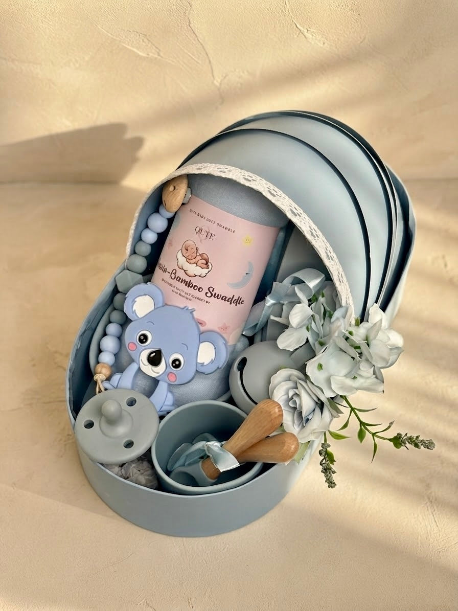 Luxury baby boy newborn gift hamper with silicone feeding set in a sky blue cradle-shaped box by Qute Qatar, featuring a bamboo swaddle, koala teether, silicone cup, and wooden accessories for delivery in Doha.