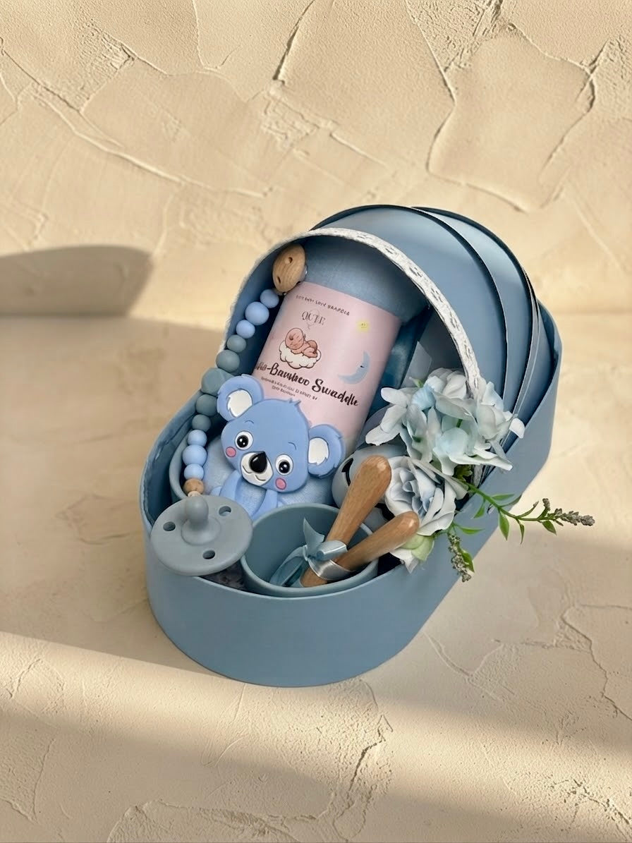 Luxury baby boy newborn gift hamper with silicone feeding set in a sky blue cradle-shaped box by Qute Qatar, featuring a bamboo swaddle, koala teether, silicone cup, and wooden accessories for delivery in Doha.