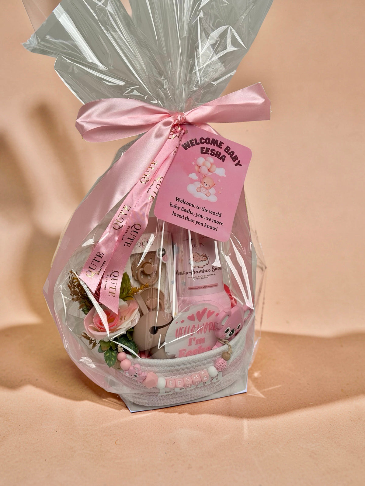 Luxury personalized baby girl gift basket wrapped in cellophane with a pink satin bow and "Welcome Baby Eesha" tag by Qute Qatar, featuring a custom woven basket, pink swaddle, koala teether, and floral accents for delivery in Doha.