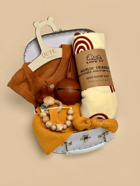 Luxury newborn baby gift set in a white keepsake suitcase by Qute Qatar, featuring a 3-6 months rust and mustard outfit, rainbow muslin swaddle, pacifiers, and wooden accessories for delivery in Doha.