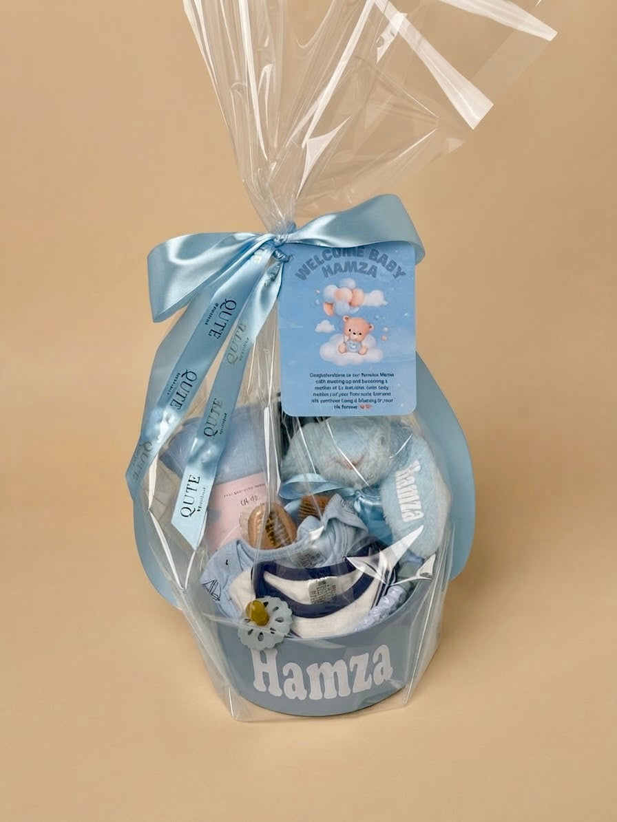 Personalized luxury newborn baby boy gift box in blue by Qute Qatar, featuring a custom keepsake suitcase named "Hamza," blue plush bunny, baby apparel, brush set, and swaddle for delivery in Doha.