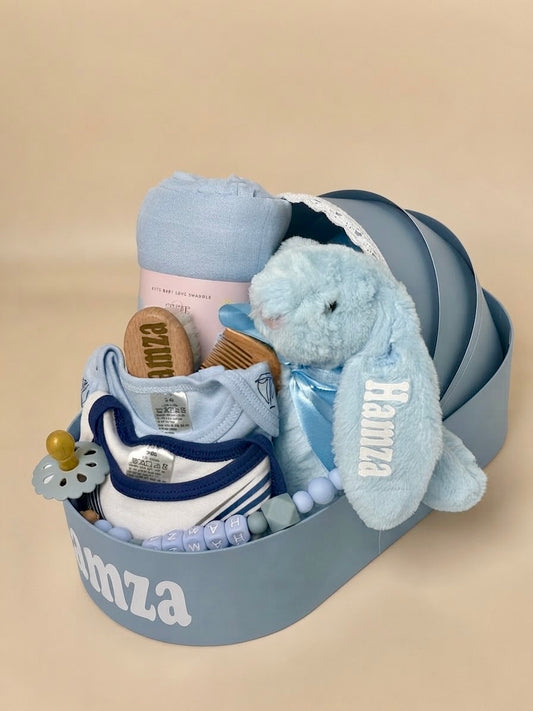 Personalized luxury newborn baby boy gift box in blue by Qute Qatar, featuring a custom keepsake suitcase named "Hamza," blue plush bunny, baby apparel, brush set, and swaddle for delivery in Doha.