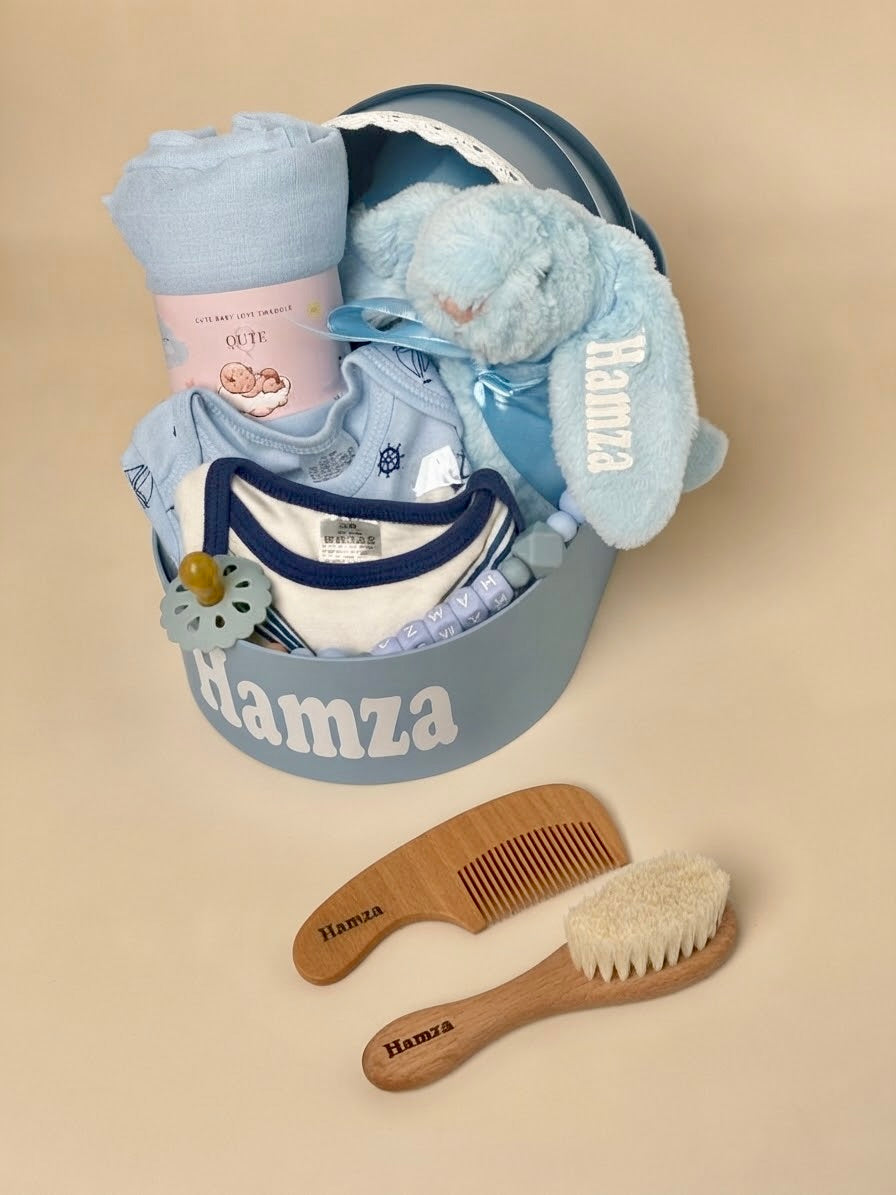 Personalized luxury newborn baby boy gift box in blue by Qute Qatar, featuring a custom keepsake suitcase named "Hamza," blue plush bunny, baby apparel, brush set, and swaddle for delivery in Doha.