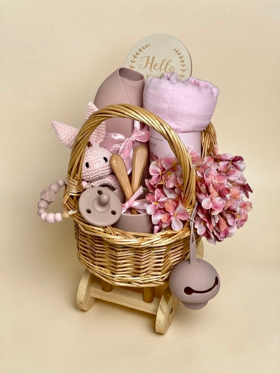 Luxury newborn baby girl feeding and blanket gift set in a wheeled wooden carriage basket by Qute Qatar, featuring a pink silicone feeding set, crochet rattle, baby blanket, and floral accents for delivery in Doha.  