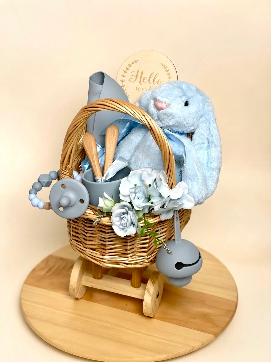 Luxury newborn baby boy feeding gift set in a wheeled wooden carriage basket by Qute Qatar, featuring a blue silicone feeding set, plush bunny, milestone disc, and floral accents for delivery in Doha.  